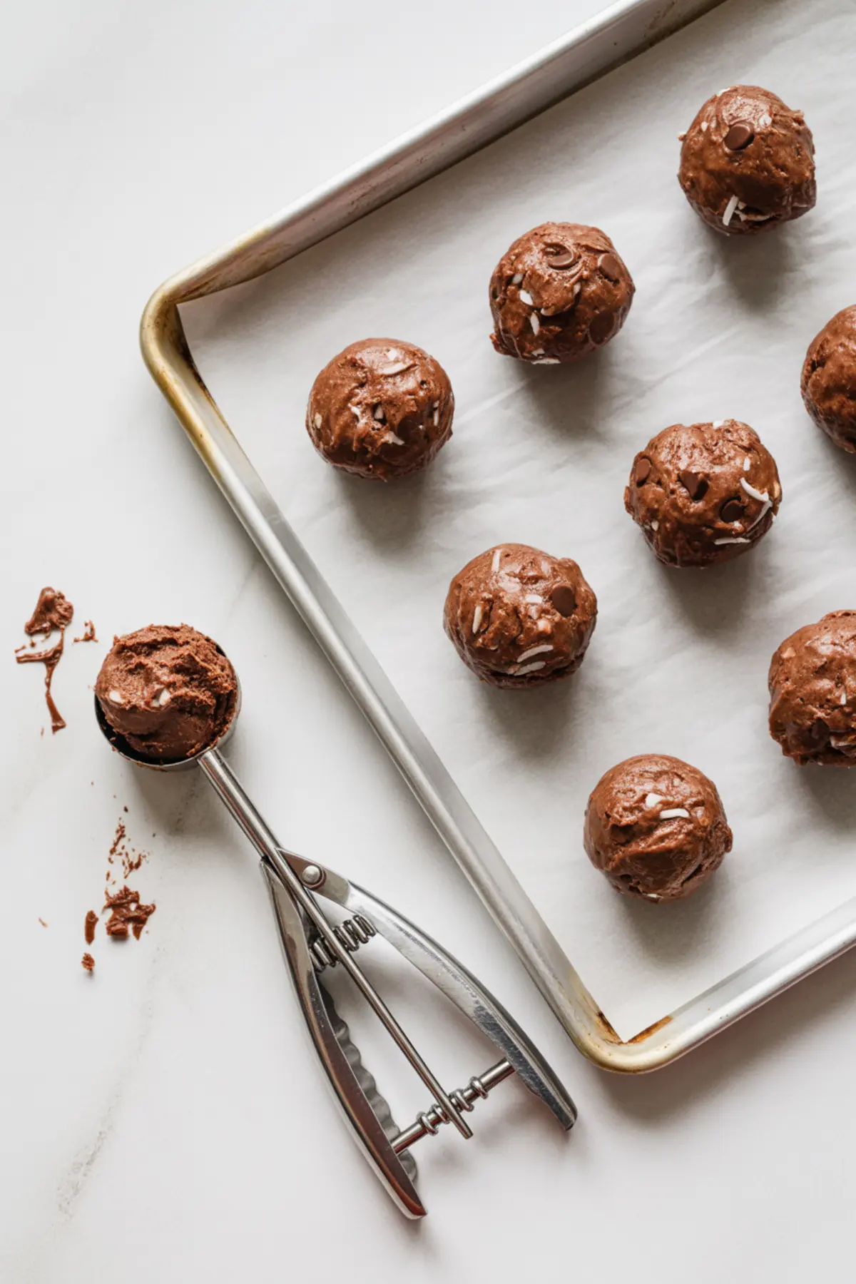 Scoops of chocolate coconut cookie dough arranged on a parchment-lined baking sheet, with a metal cookie scoop beside it.
