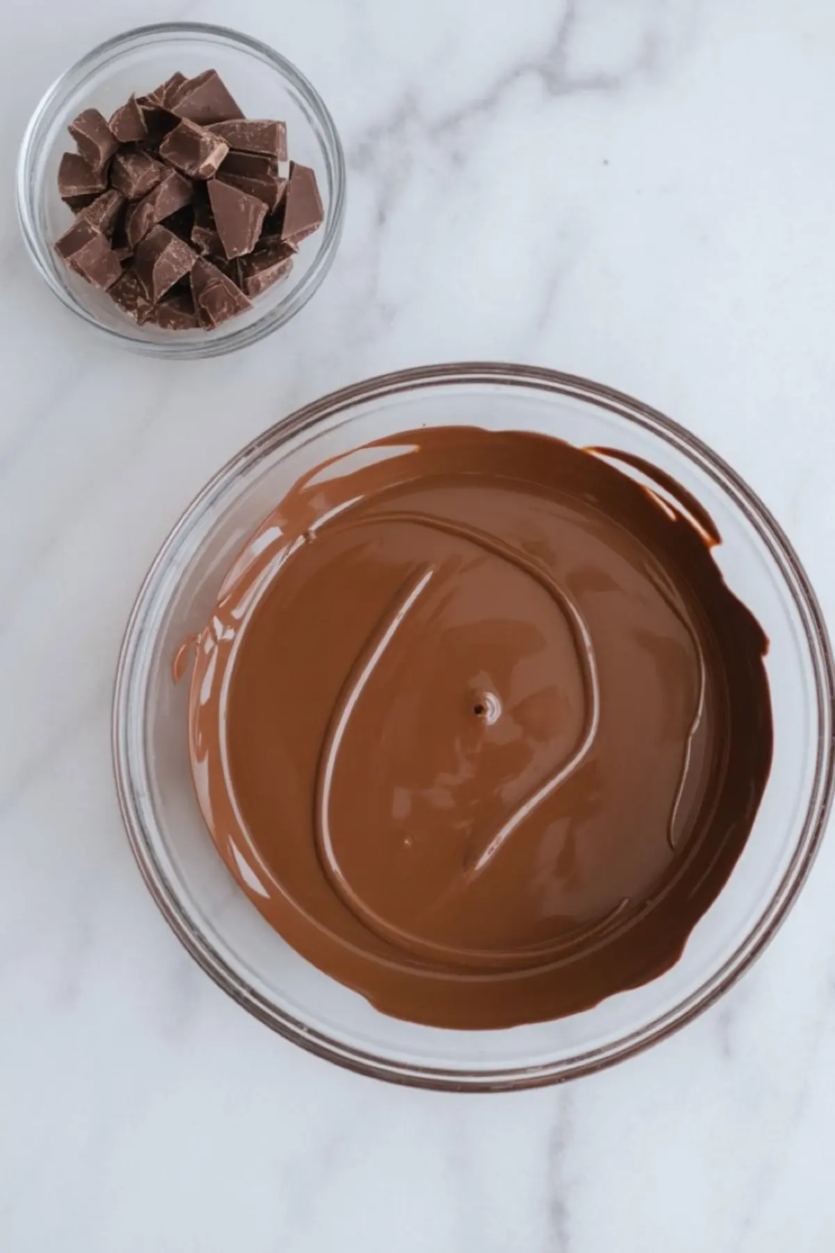 A bowl of melted milk chocolate next to a smaller bowl filled with chopped chocolate pieces on a white marble background.
