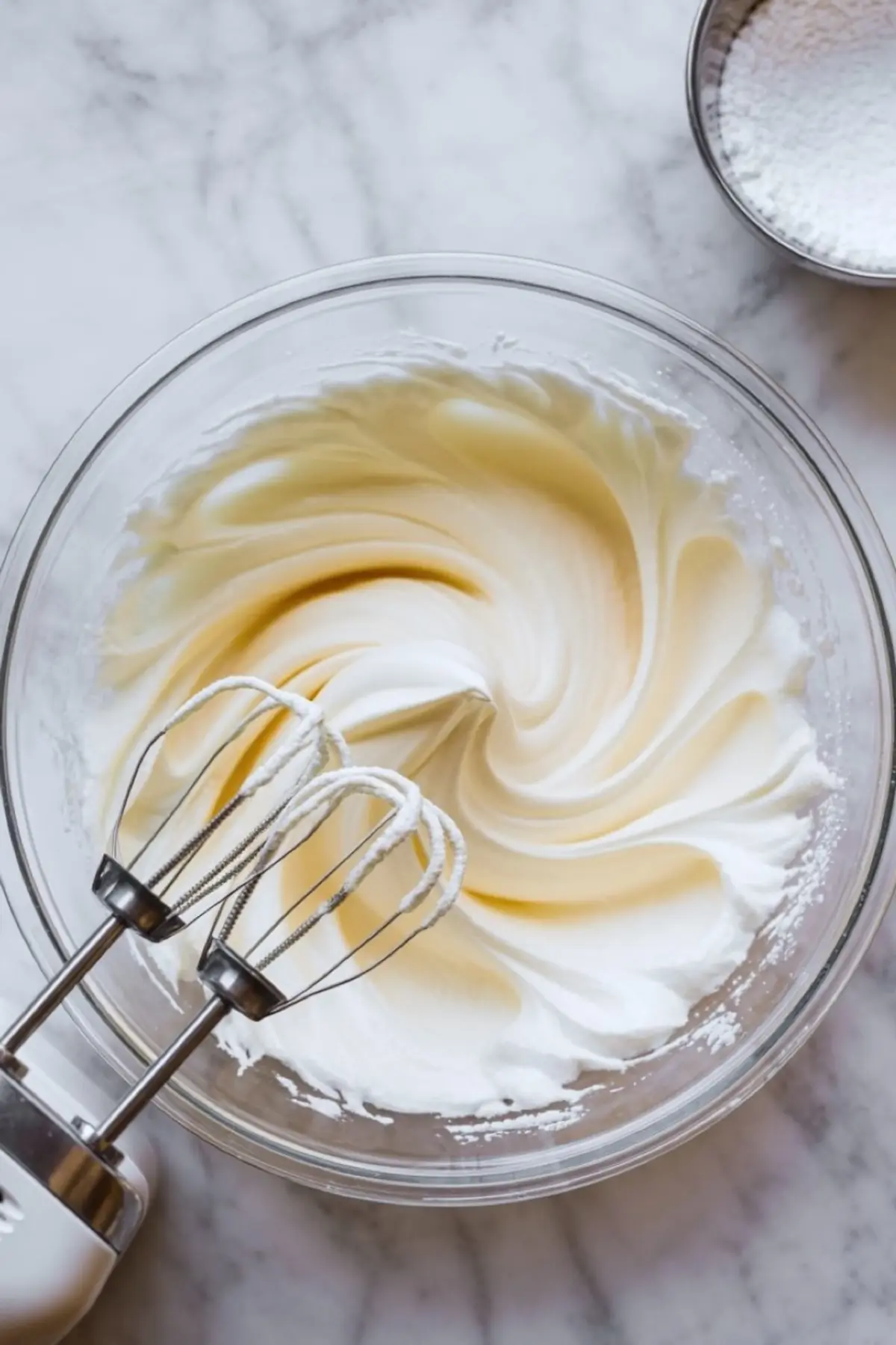 A bowl of whipped cream with soft peaks on a marble surface, featuring two hand mixer beaters resting inside.
