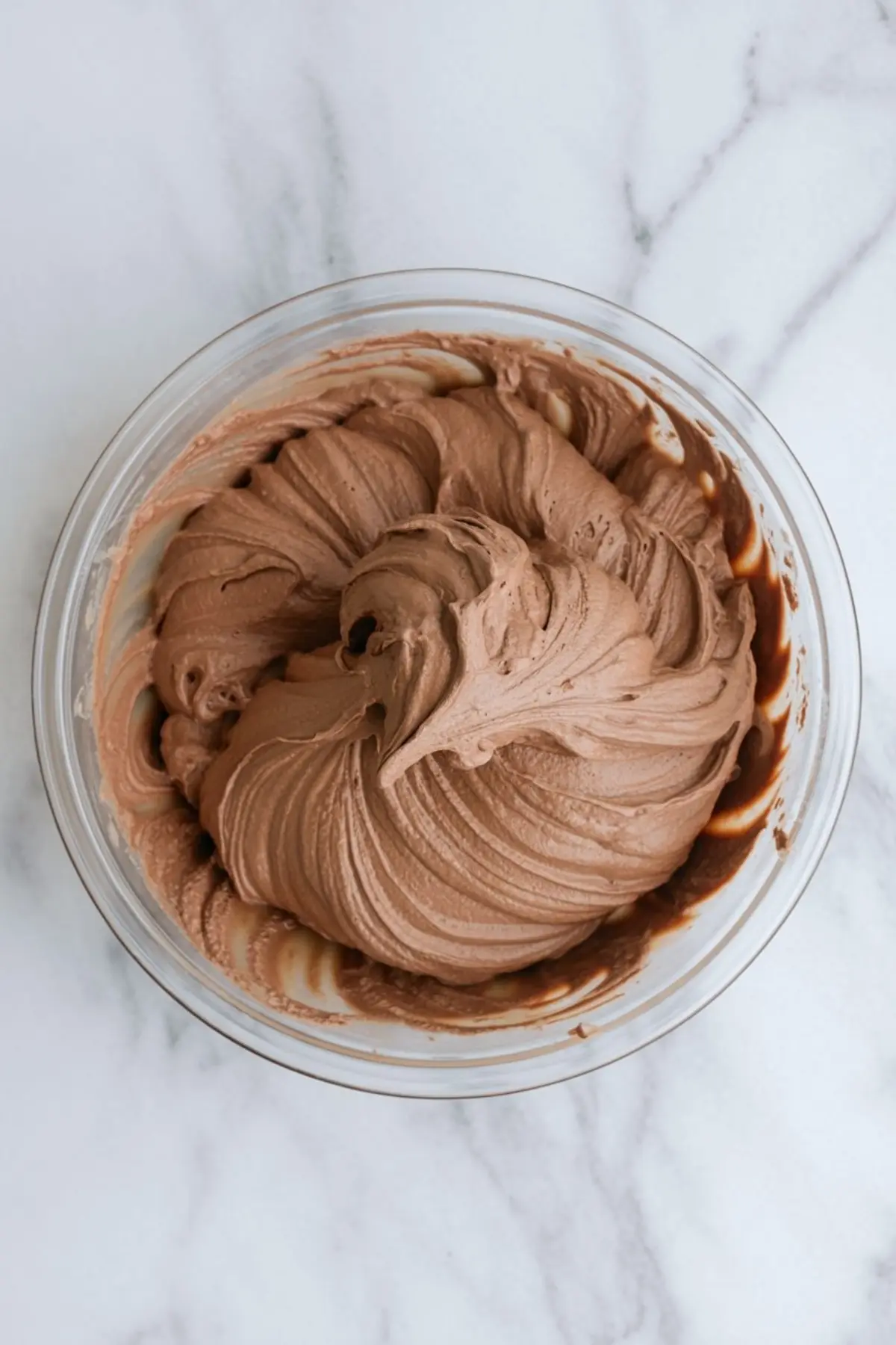 A fully mixed batch of chocolate mousse with a smooth, thick texture in a glass bowl on a marble surface, featuring swirls from a spatula.
