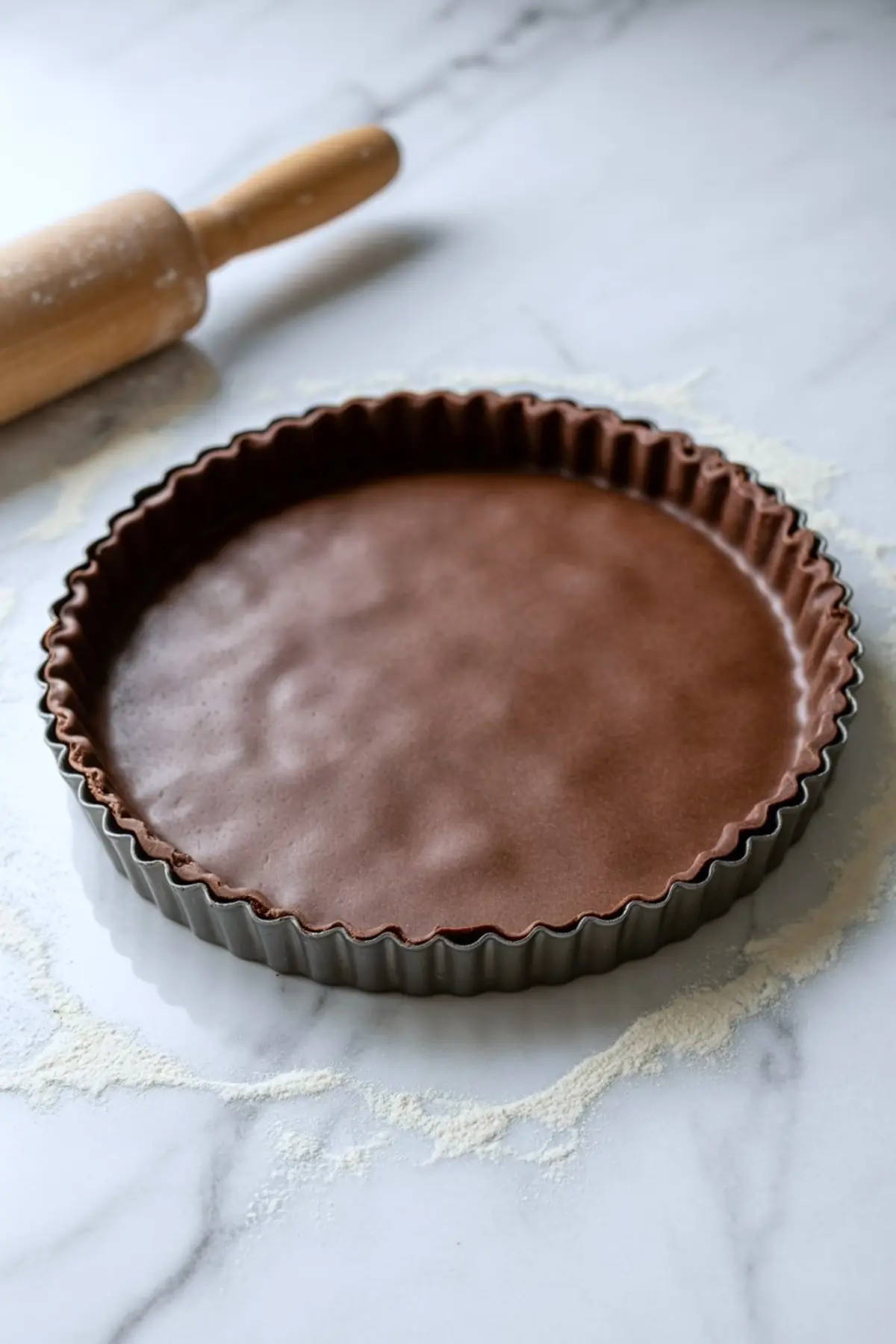 Chocolate tart crust pressed into a fluted tart pan, resting on a marble surface with scattered flour and a wooden rolling pin in the background
