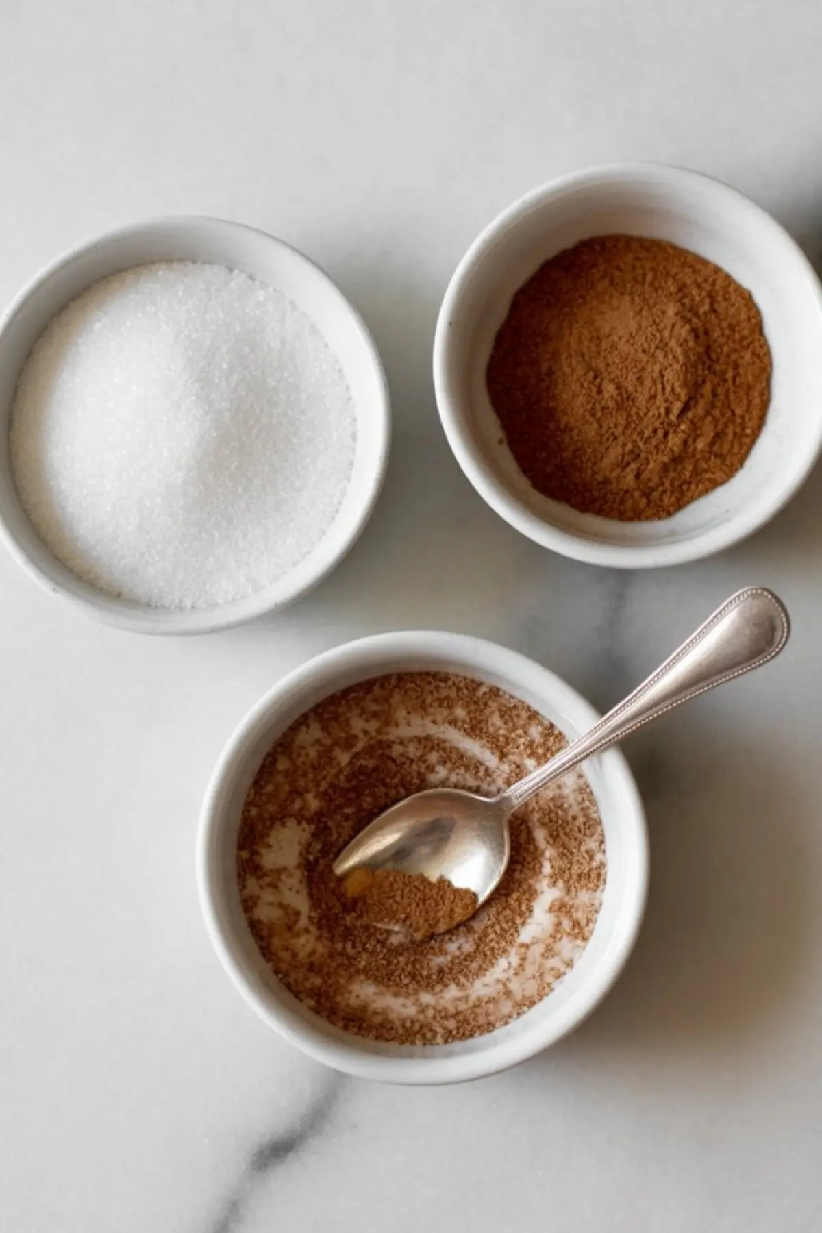 Three small white bowls containing white granulated sugar, cinnamon powder, and a cinnamon-sugar blend with a silver spoon, placed on a white marble surface.
