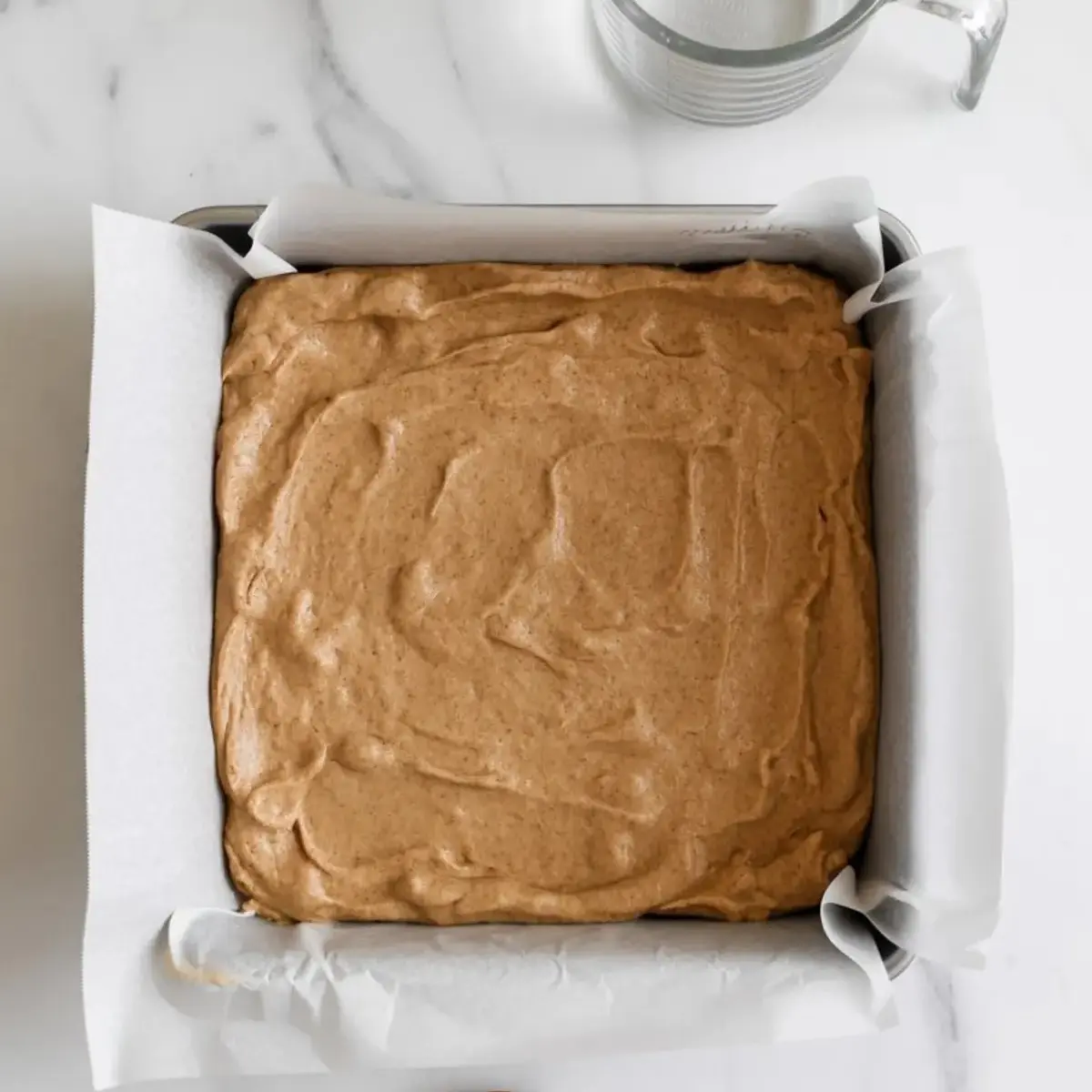 A square metal baking pan lined with parchment paper and filled with a layer of cinnamon-sugar dough, next to a glass measuring cup and wooden spoon on a marble counter.
