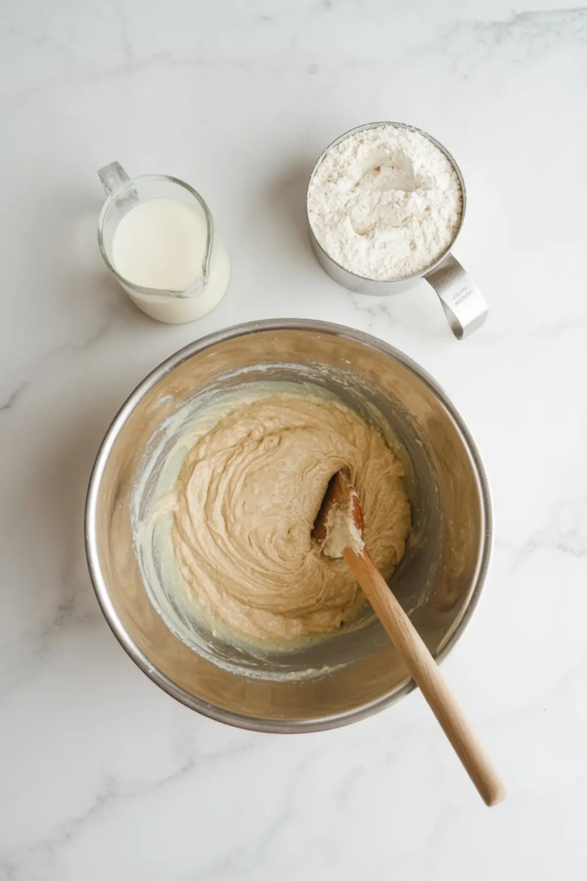 Smooth cupcake batter in a metal bowl with a wooden spatula, placed beside a glass of milk and a metal measuring cup filled with flour on a marble surface.
