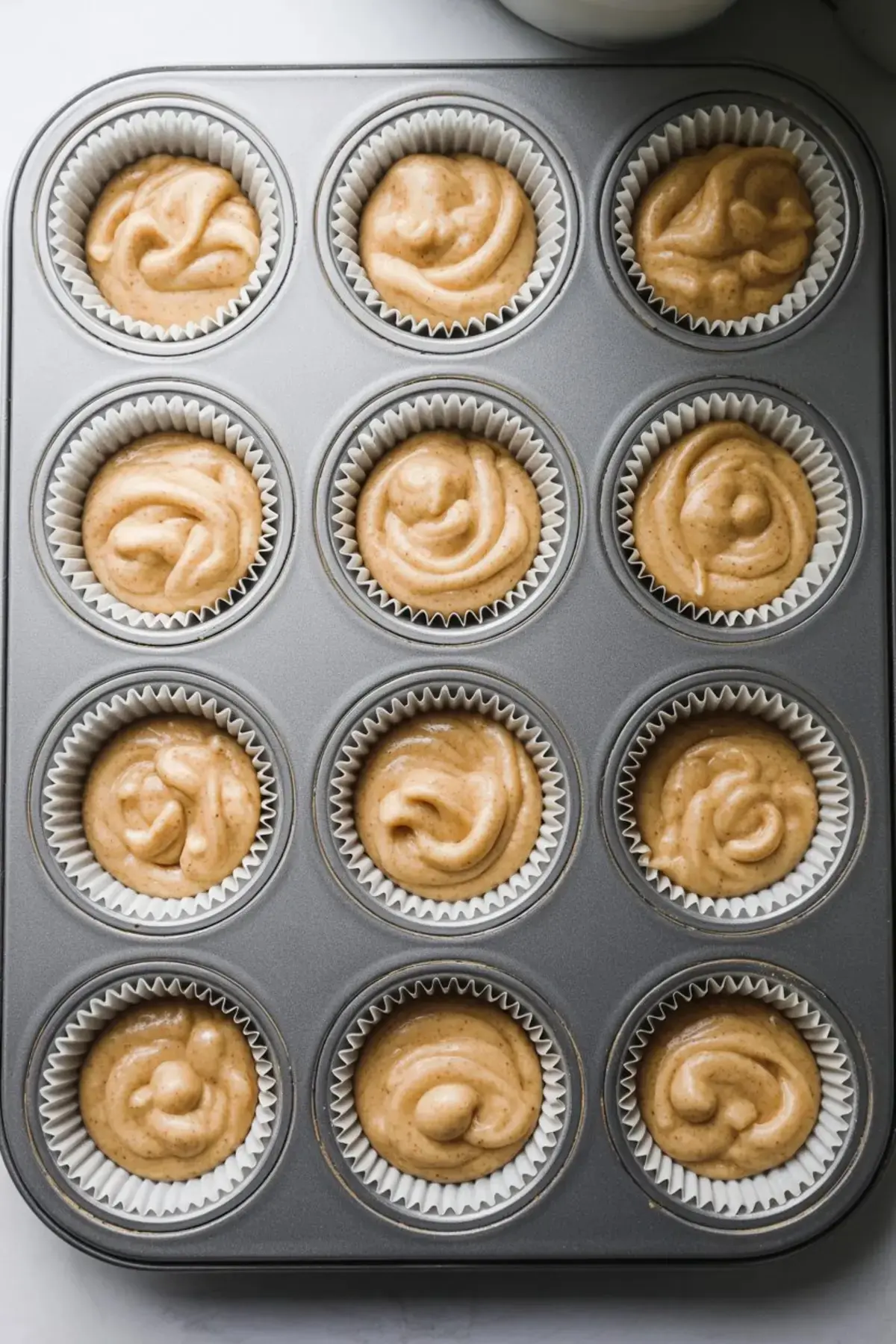 Cupcake batter portioned into a 12-cup muffin tin lined with white paper liners, ready for baking, showcasing a cinnamon-speckled mixture.
