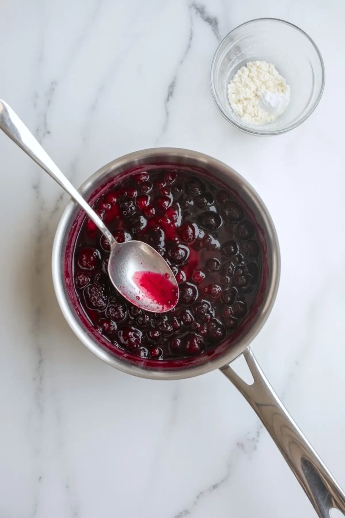 Saucepan filled with glossy cherry compote made from whole berries in a thick red syrup, placed on a marble counter with a spoon and a small bowl of cornstarch beside it.