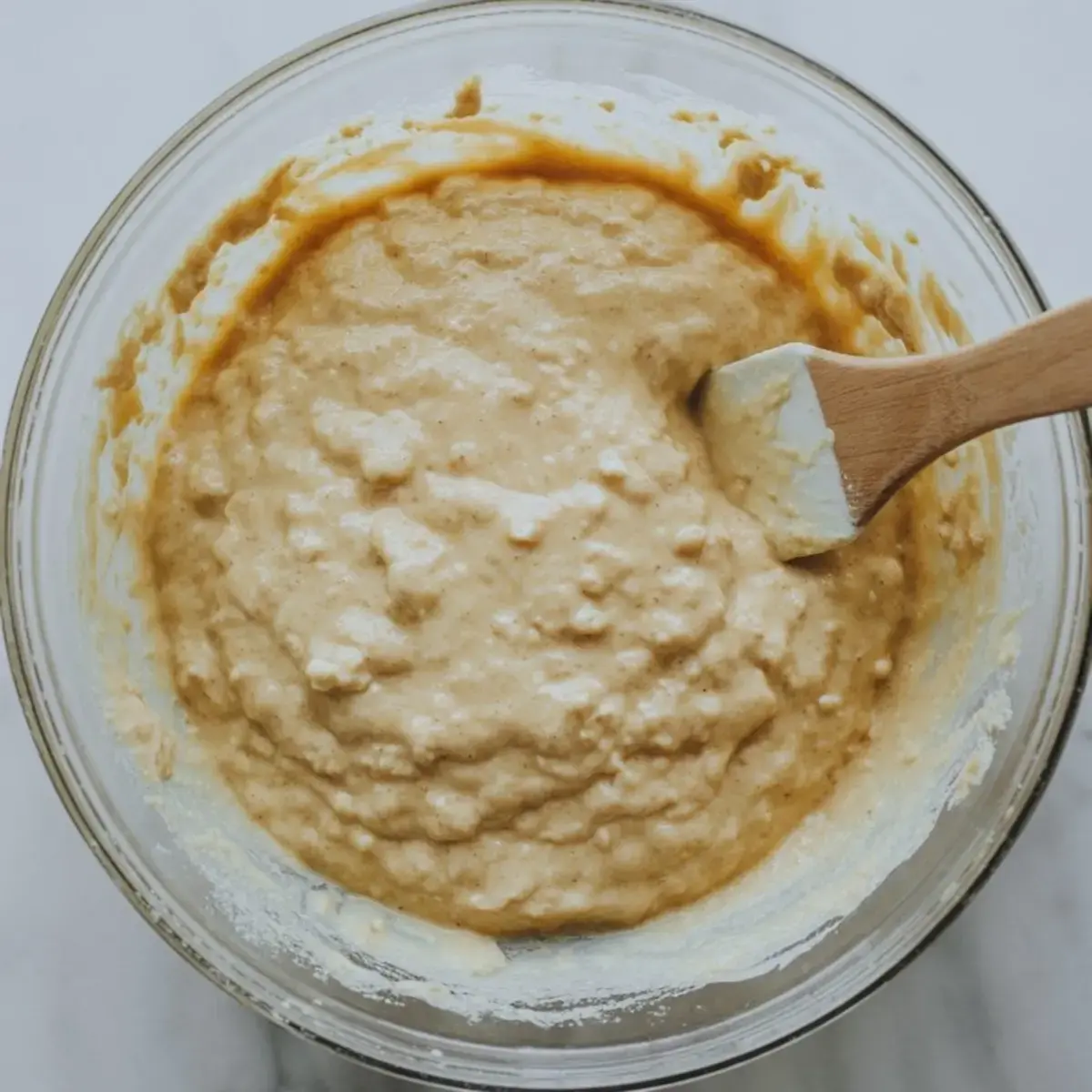 Glass bowl filled with thick cottage cheese pancake batter being stirred with a spatula, placed on a light marble surface.
