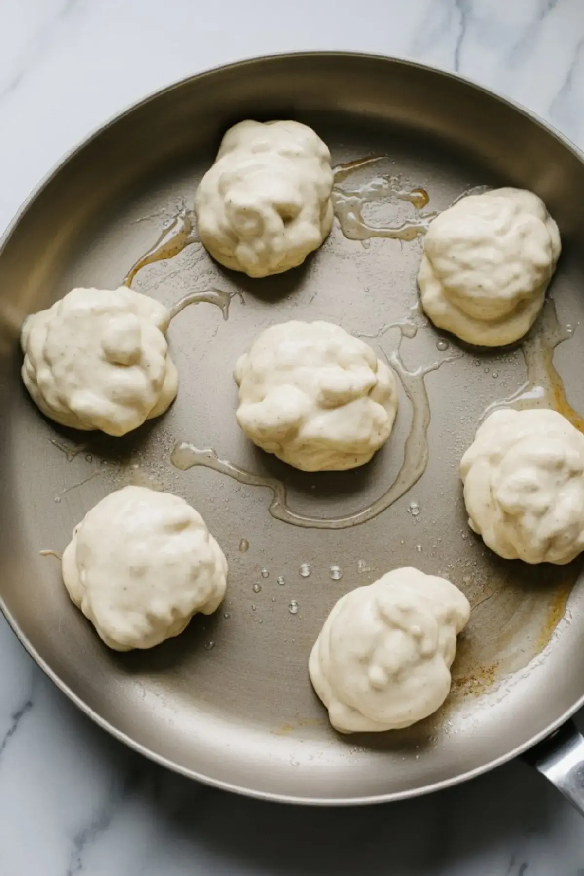Raw cottage cheese pancake batter dropped in dollops onto a greased frying pan, ready for cooking on a stovetop.
