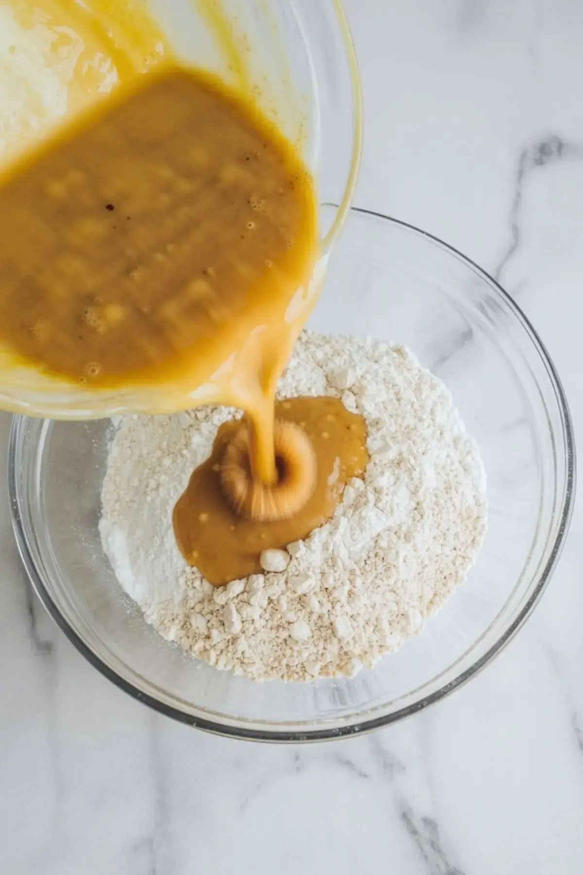 Glass bowl of dry pancake mix with a thick stream of wet batter being poured in, highlighting the process of combining ingredients.

