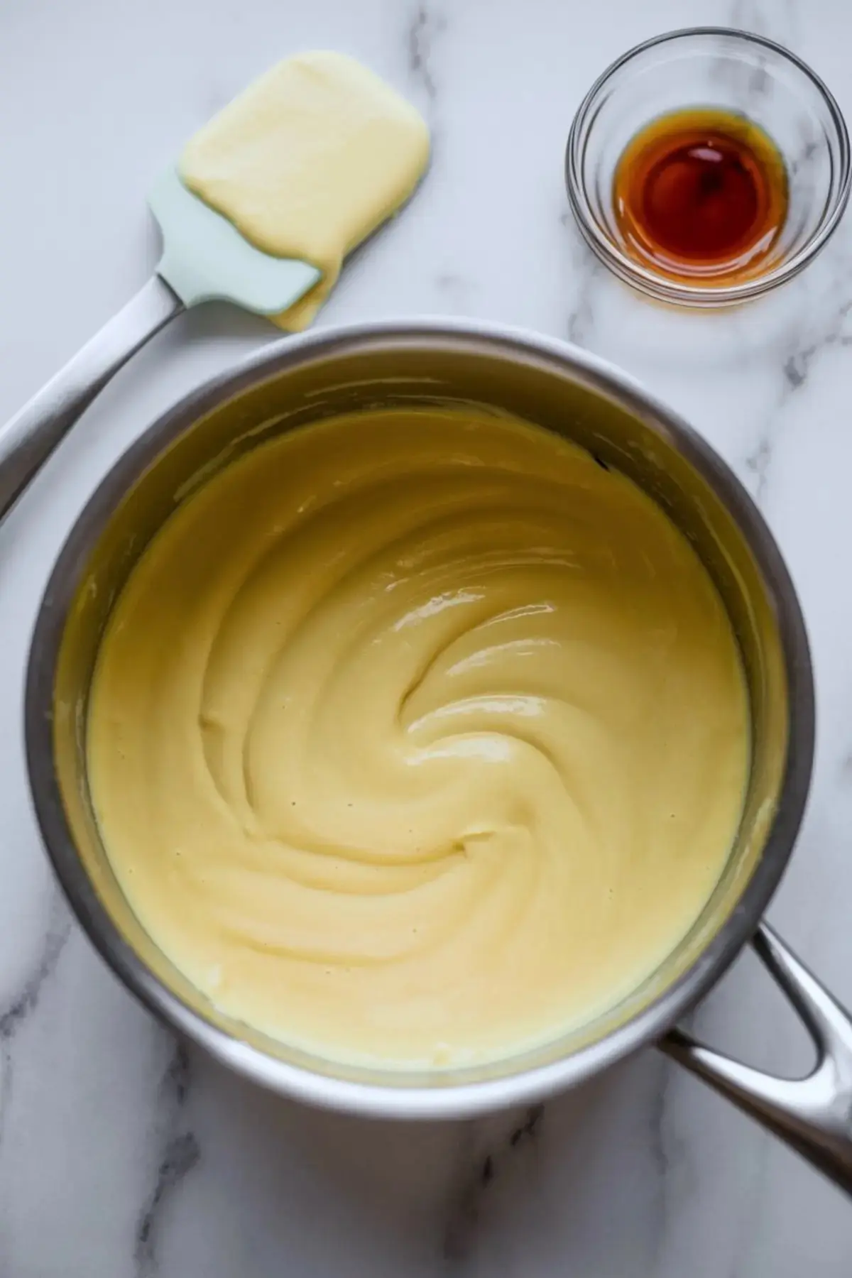 Silky vanilla pastry cream fills a stainless steel saucepan, with a green spatula coated in cream and a glass bowl of vanilla extract arranged beside it on a marble background.