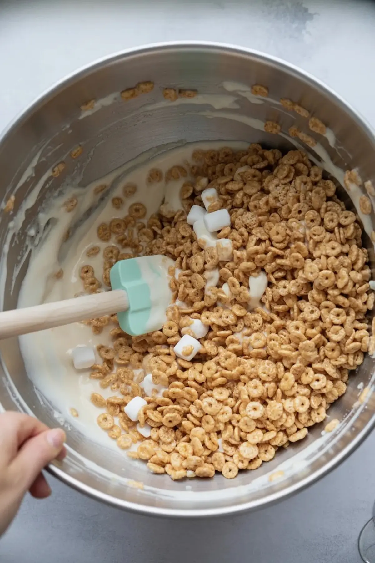 Large mixing bowl filled with honey nut cereal and mini marshmallows being stirred with a mint green spatula into a creamy melted marshmallow mixture.