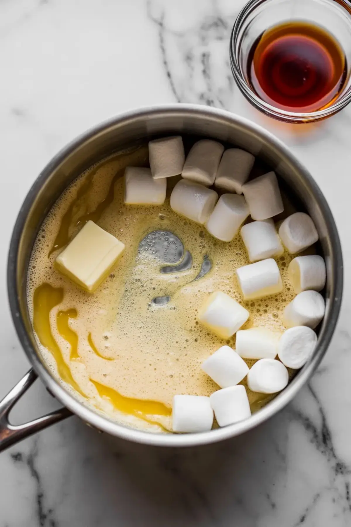 Stainless steel saucepan containing melting butter, mini marshmallows, and light brown sugar mixture, with a small bowl of vanilla extract beside it on a marble countertop.
