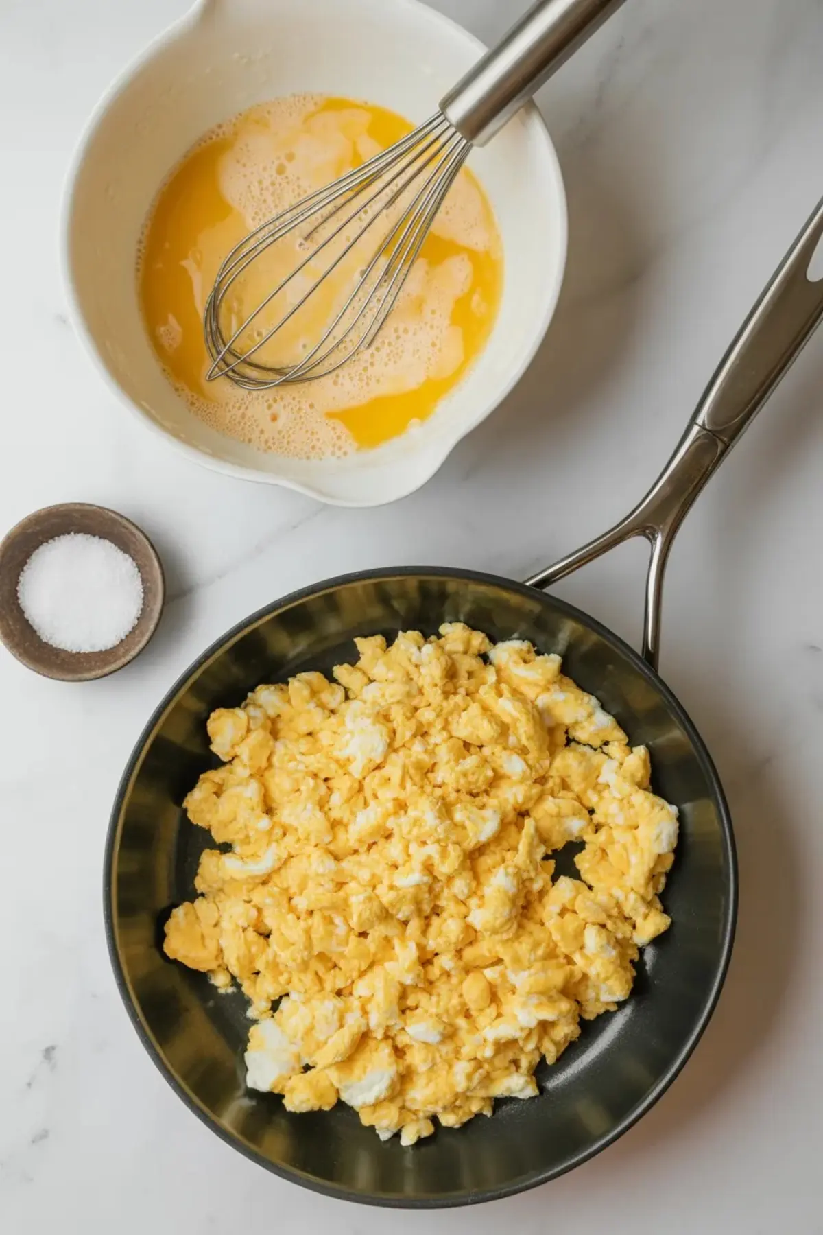 Overhead view of fluffy scrambled eggs in a nonstick skillet, next to a mixing bowl with beaten eggs and whisk, and a small dish of coarse salt on a marble countertop.