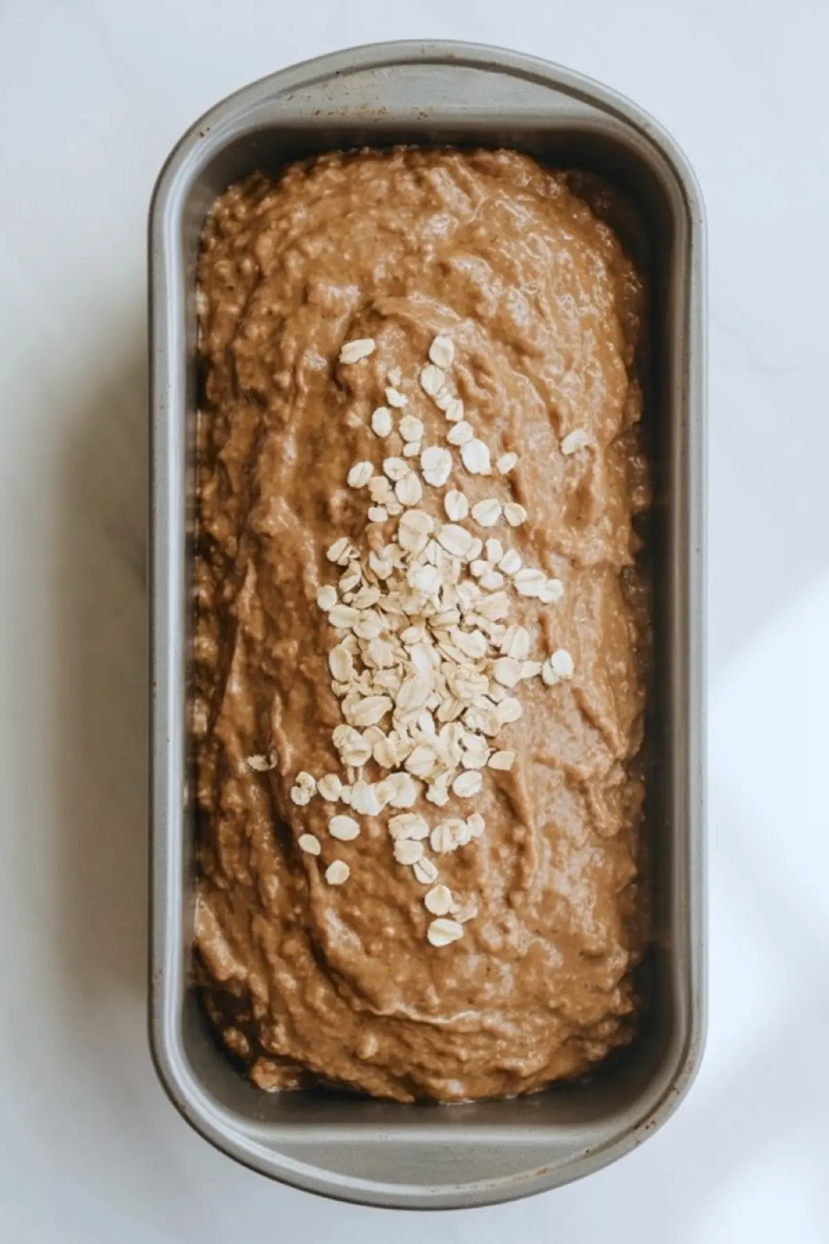 Unbaked Guinness oatmeal bread batter in a metal loaf pan topped with raw oats, ready for baking, placed on a light background for a clean and rustic aesthetic.
