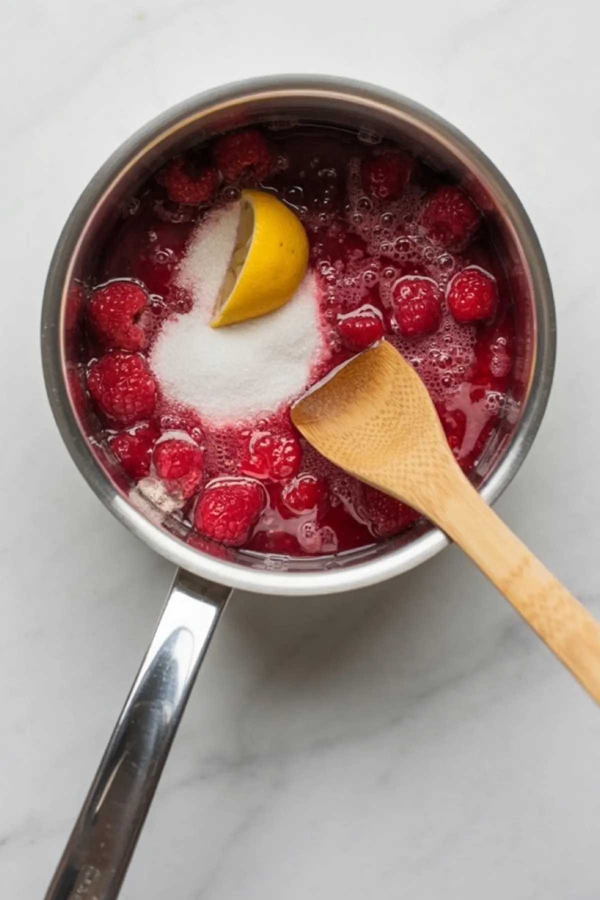 Fresh raspberries, granulated sugar, and a lemon wedge are combined in a saucepan for a homemade raspberry sauce, with a wooden spatula resting on the edge.
