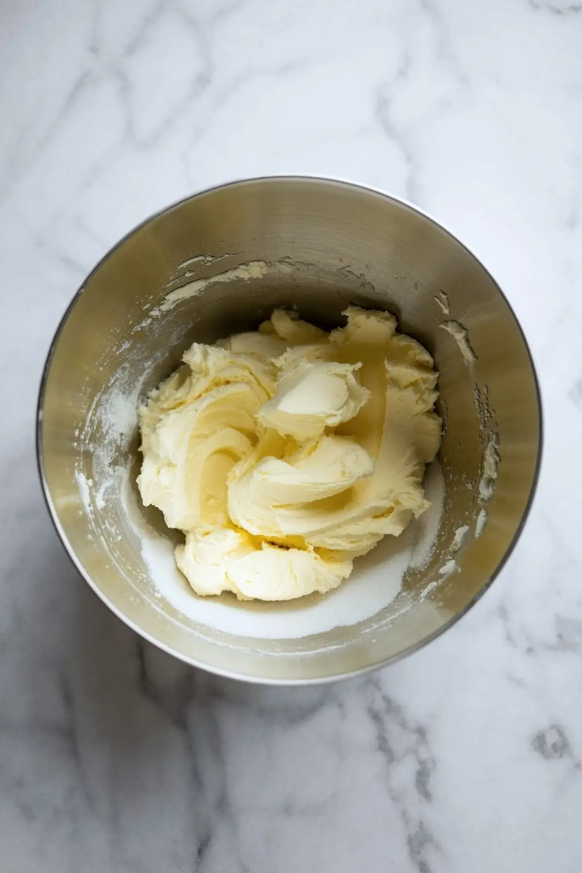 Creamed butter and sugar mixture in a stainless steel mixing bowl on a marble countertop, showing a fluffy and smooth texture ideal for baking cupcakes or cookies.
