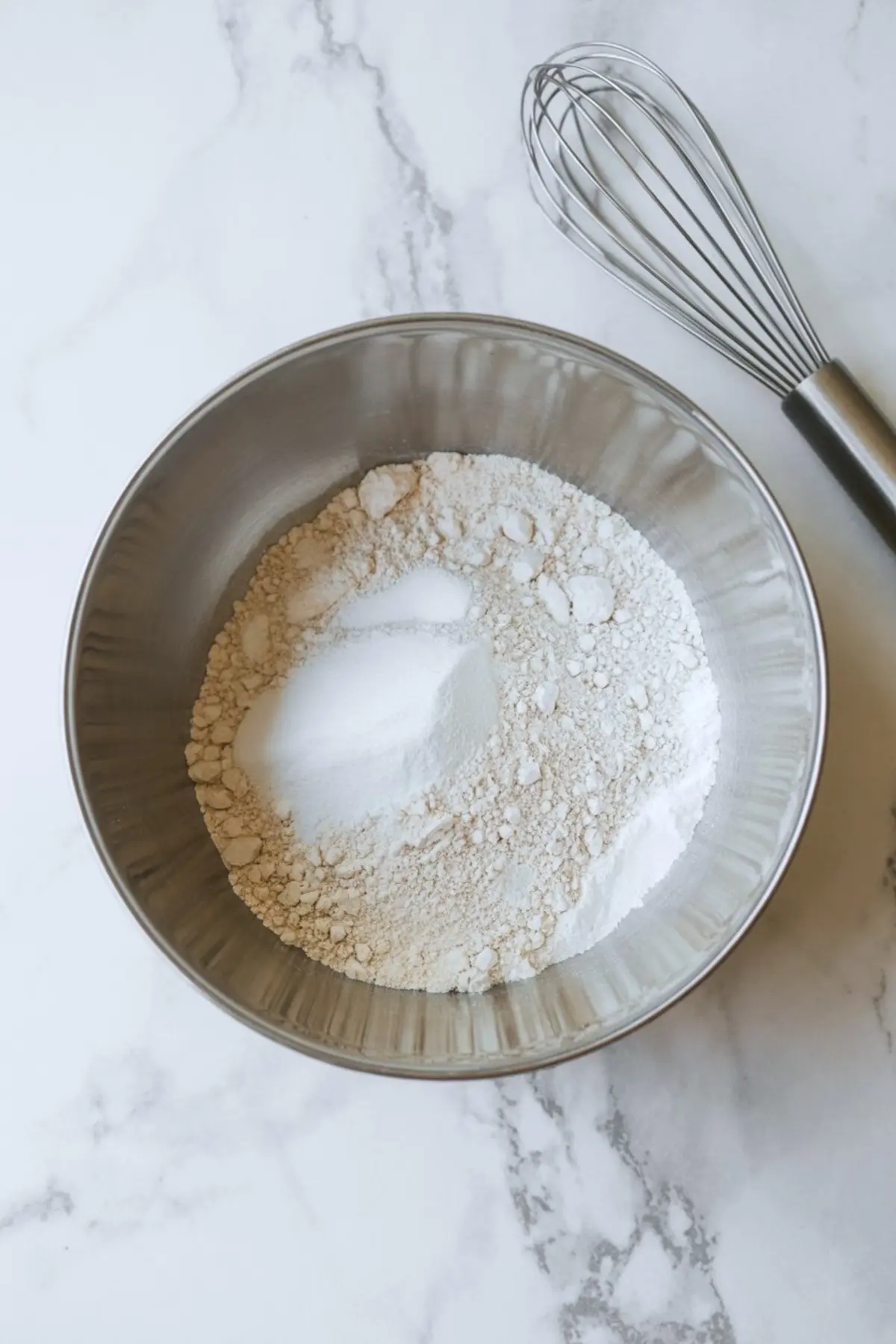 Dry ingredients including flour, baking powder, and salt in a metal mixing bowl beside a metal whisk, set on a white marble counter for cupcake preparation.
