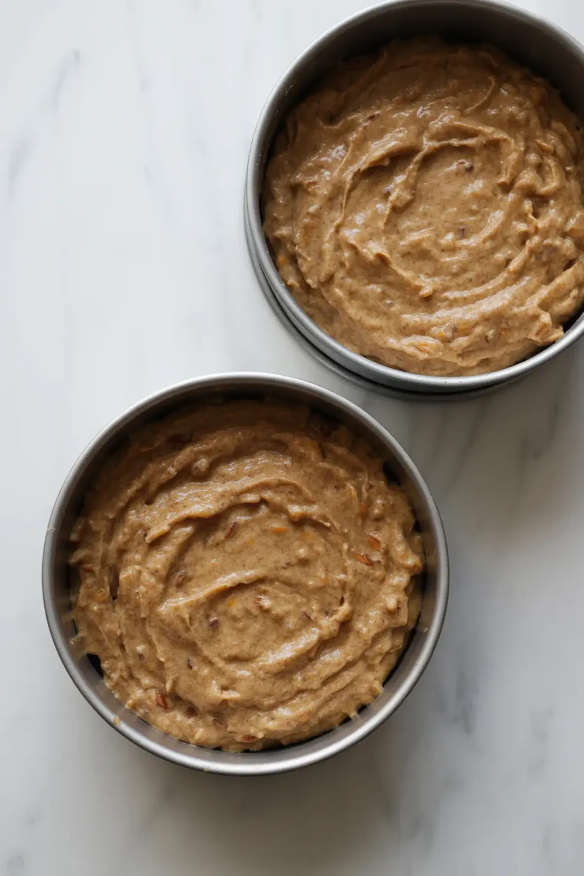 Two round cake pans filled with unbaked carrot cake batter speckled with nuts and grated carrots, placed side by side on a white marble counter.
