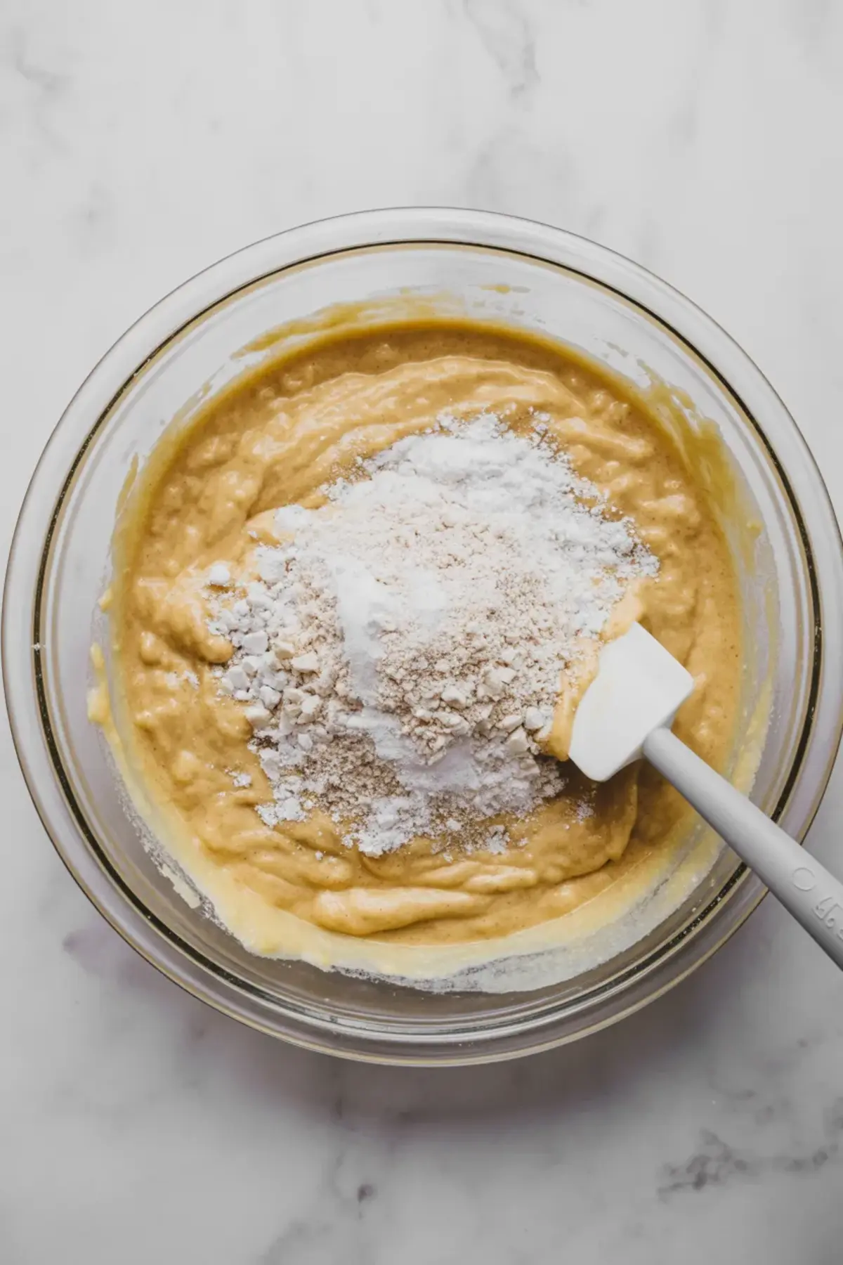 Glass mixing bowl filled with thick carrot cake batter topped with oat flour and baking powder, with a white spatula partially blending the mixture on a marble surface.

