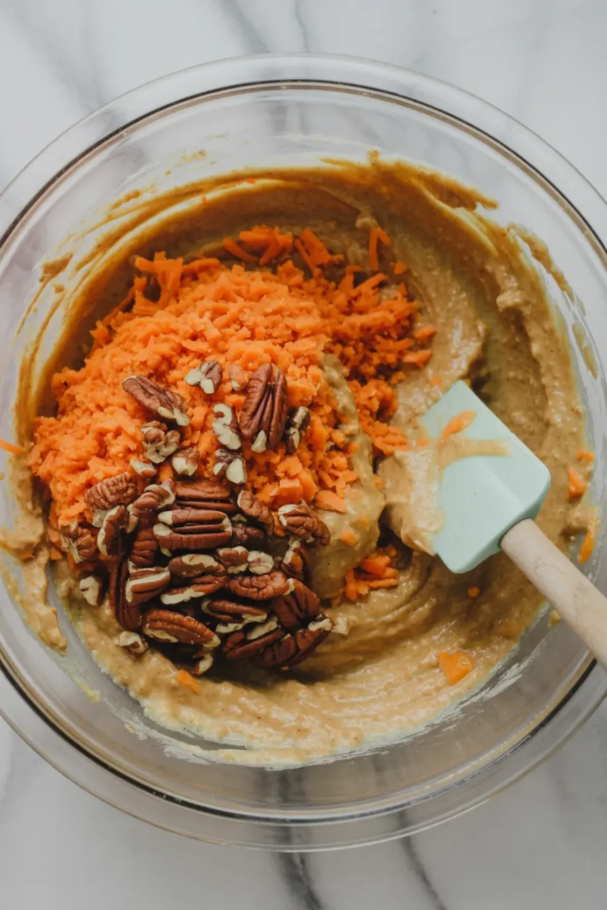 Clear bowl containing carrot cake batter with freshly grated carrots and raw pecan halves on top, next to a teal spatula ready to mix on a marble surface.
