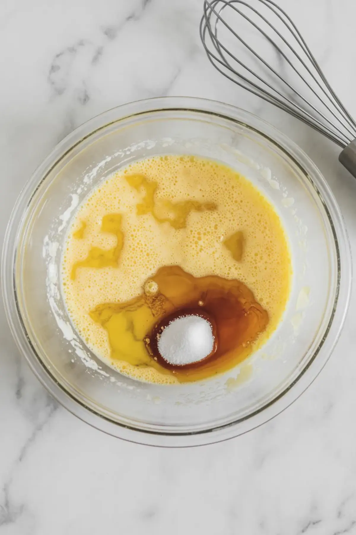 Glass bowl with whisked wet ingredients including eggs, vanilla extract, oil, and granulated sweetener, with a metal whisk placed beside it on a marble counter.
