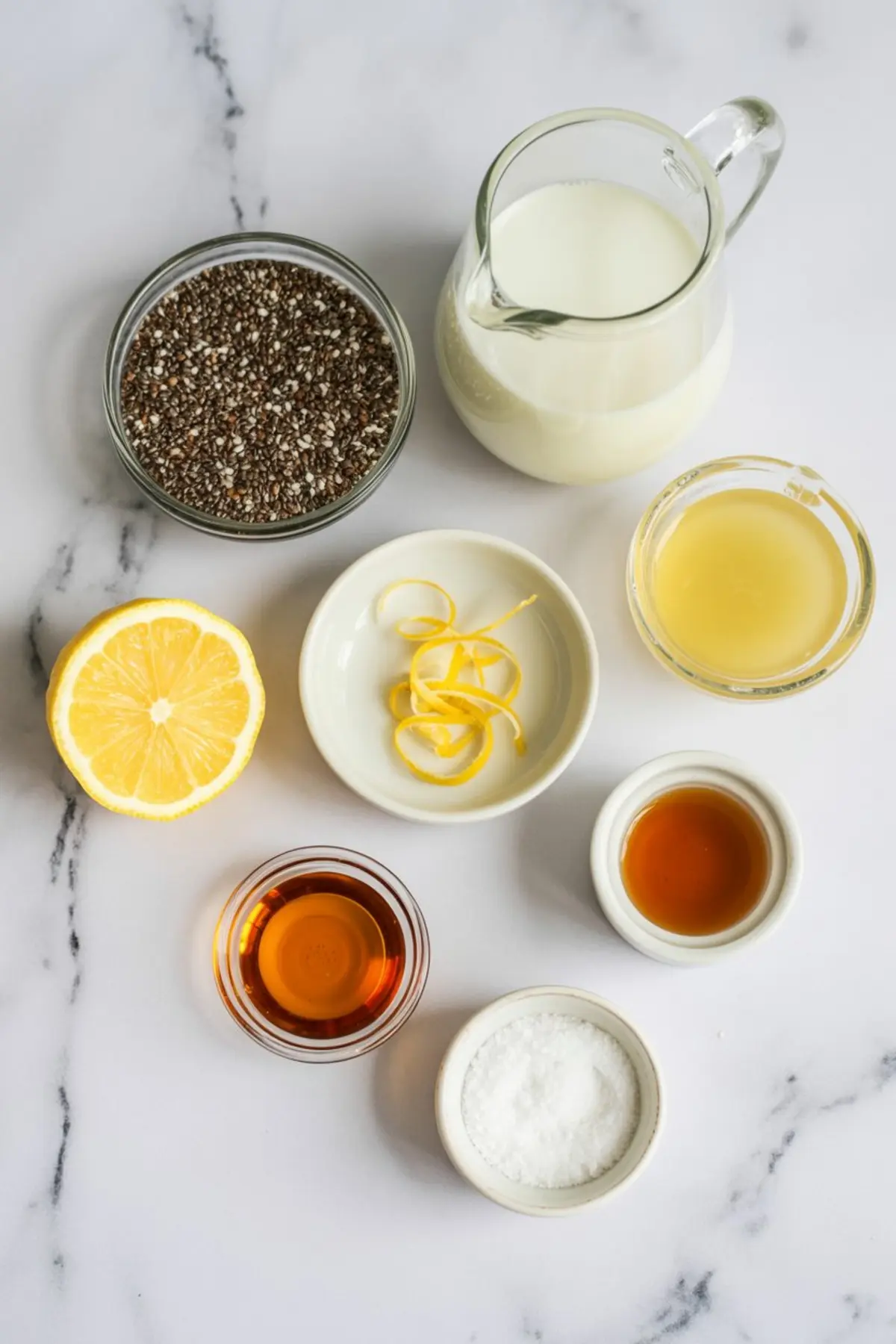 Flat lay of fresh ingredients for lemon chia seed pudding, including a halved lemon, chia seeds, milk, lemon juice, lemon zest, maple syrup, vanilla extract, and a small bowl of salt, all arranged on a white marble surface.

