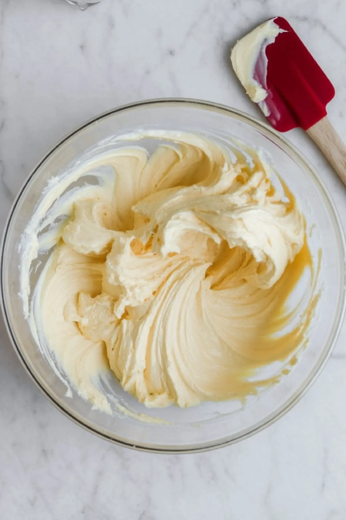 A bowl of whipped lemon cream cheese filling is shown mid-mix with a red spatula resting on the edge. The texture is light and airy, showing the rich swirl of the freshly blended cake filling ideal for citrus-based desserts.
