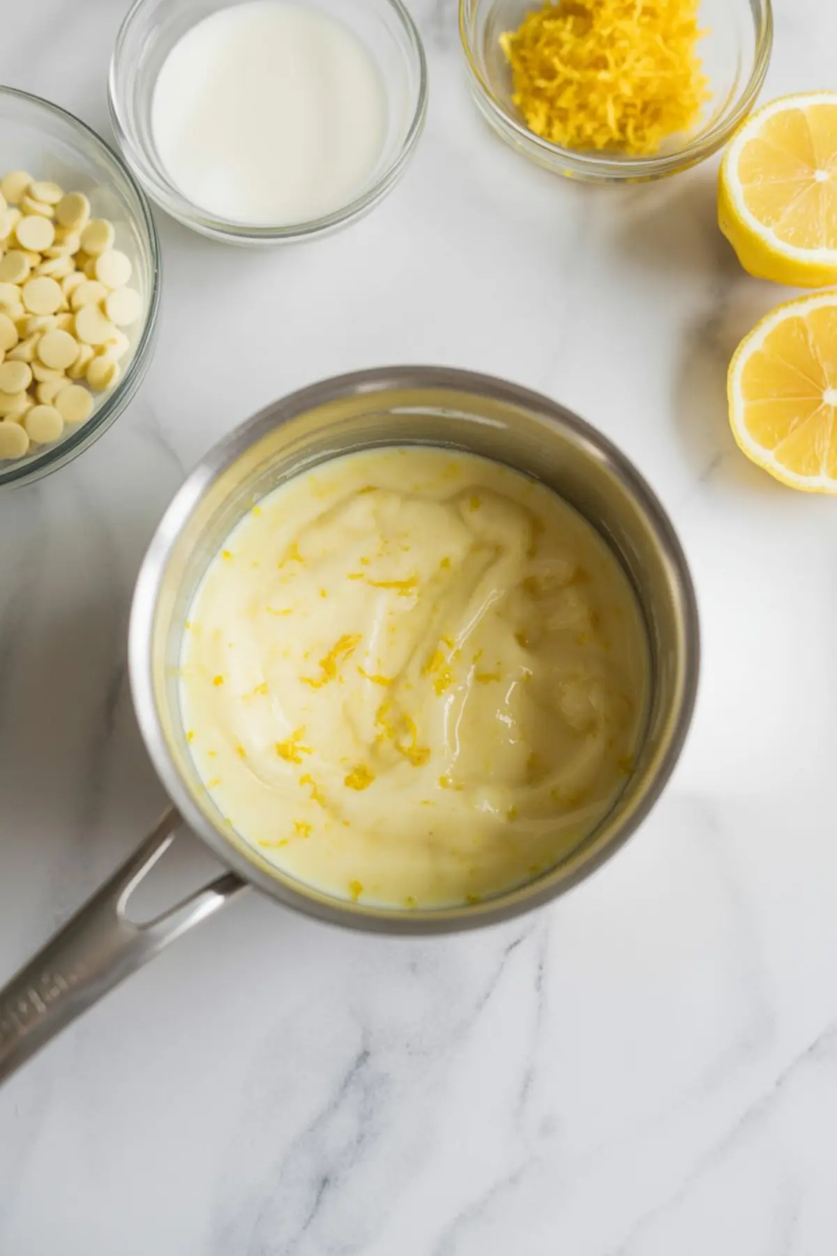 Ingredients for lemon white chocolate filling laid out on a white marble surface, including lemon zest, white chocolate chips, milk, lemon halves, and a saucepan with smooth lemon mixture.
