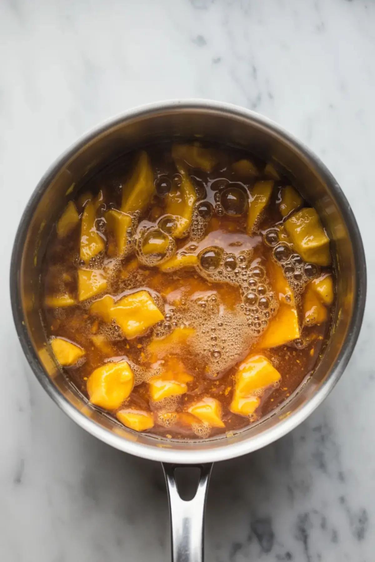 Cooked mango chutney bubbling in a stainless steel saucepan, with visible chunks of mango simmering in a thick, spiced liquid.