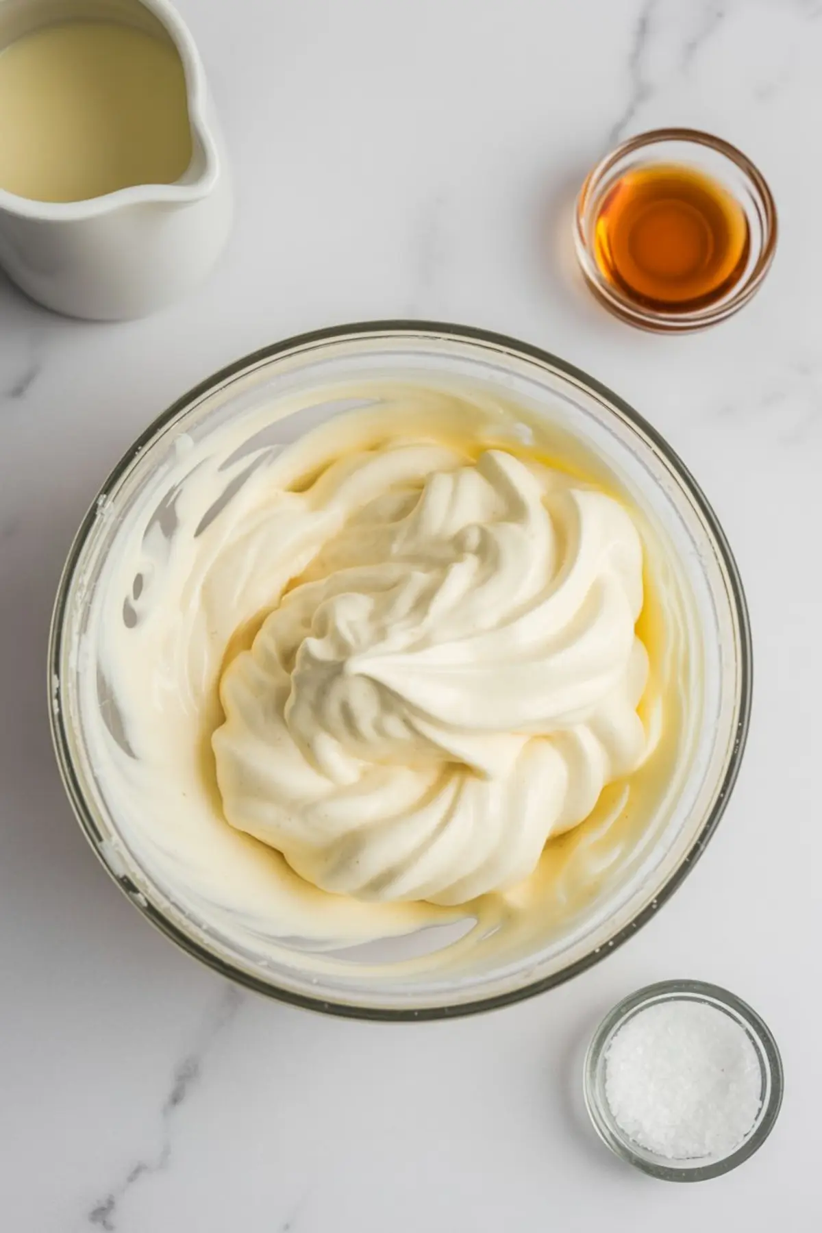 Glass bowl filled with thick whipped cream mixture, surrounded by ingredients like sweetened condensed milk, vanilla extract, and sea salt on a white marble surface.
