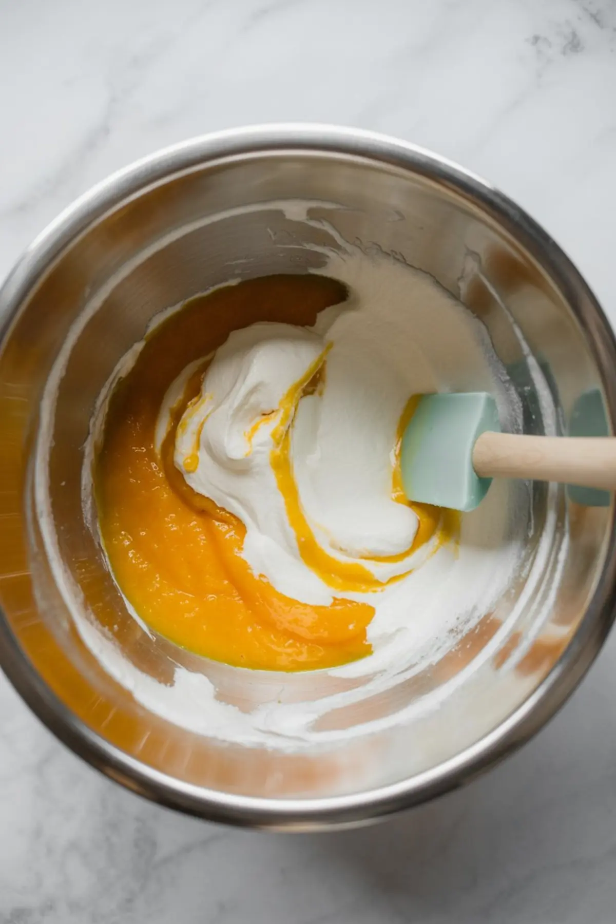 Close-up of a mixing bowl with whipped cream and mango purée being gently folded together using a silicone spatula, showing the smooth texture transition of the mousse base.
