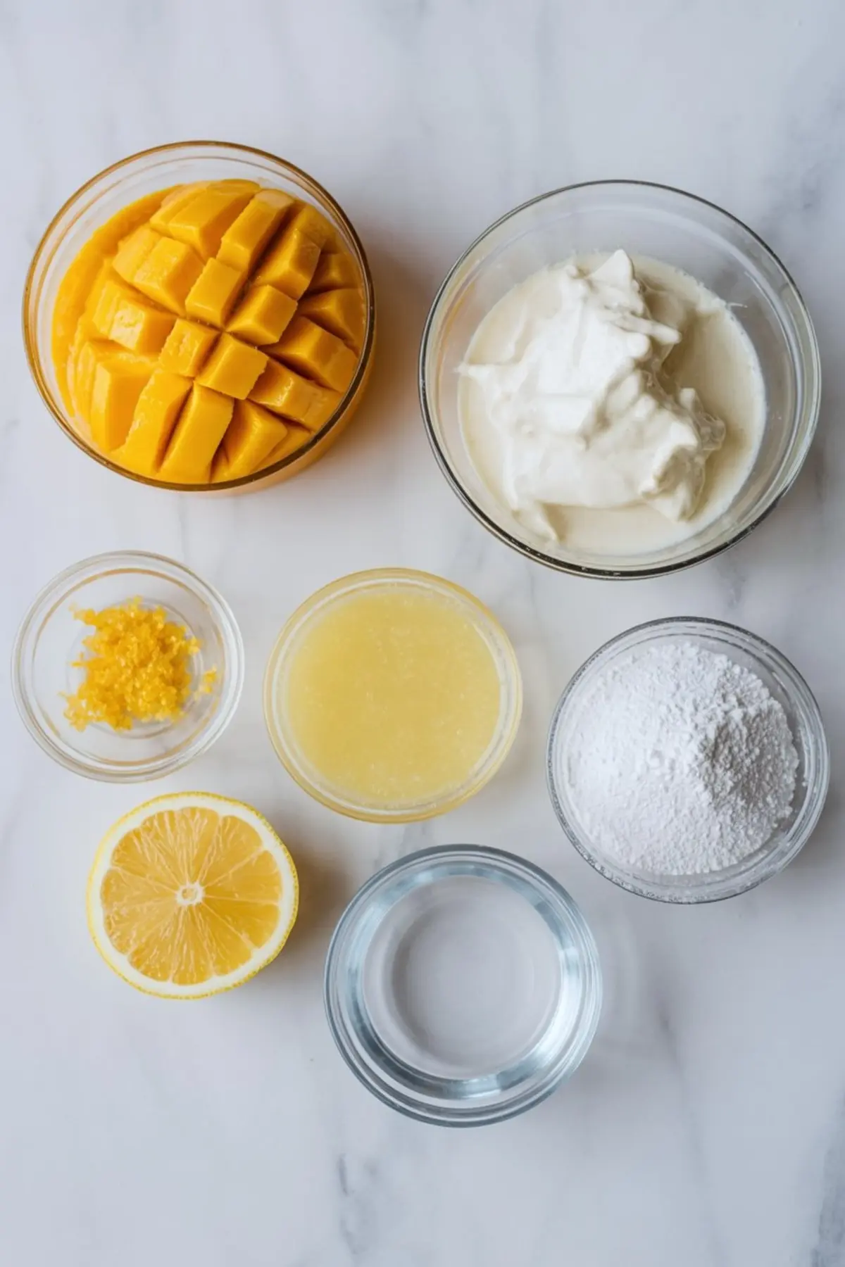 Top-down view of fresh mango mousse ingredients including cubed mango, whipped cream, powdered sugar, lemon juice, lemon zest, and water, all arranged in glass bowls on a white marble surface.
