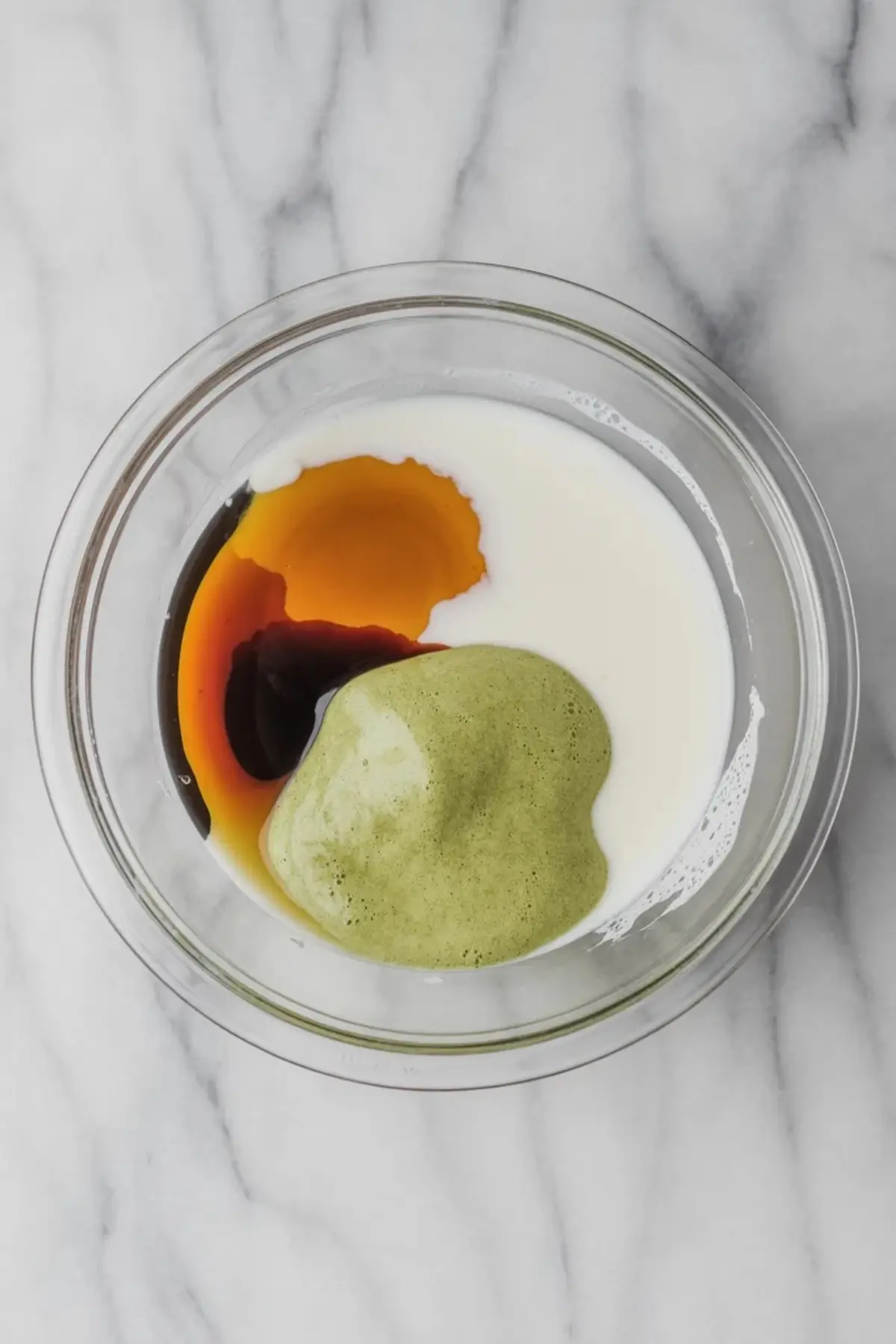 A glass mixing bowl holding coconut milk, matcha paste, and maple syrup in separate pools before mixing, placed on a light marble surface.
