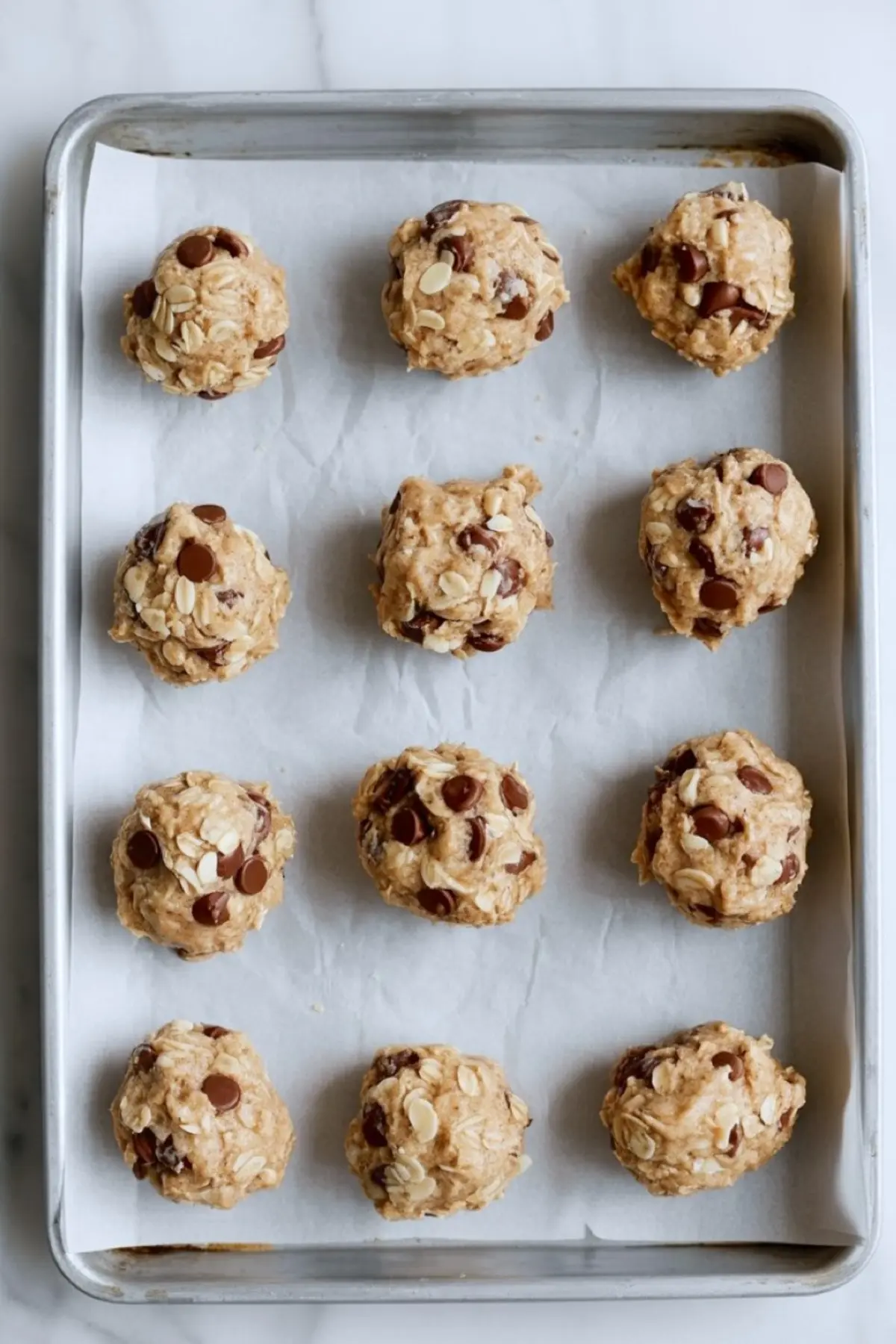 Baking sheet lined with parchment paper holding evenly spaced oatmeal chocolate chip cookie dough balls before baking.
