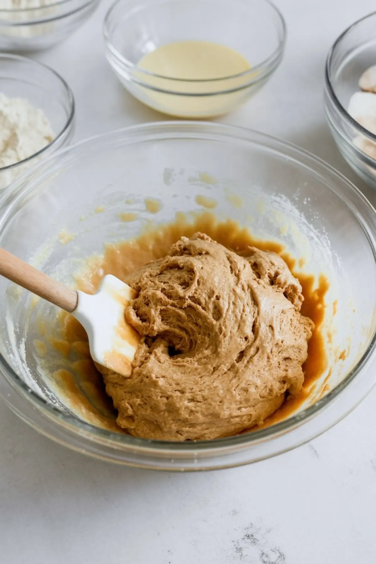 Thick cookie dough mixture in a clear glass bowl with a white spatula, surrounded by bowls of flour, sweetened condensed milk, and other ingredients on a white countertop.
