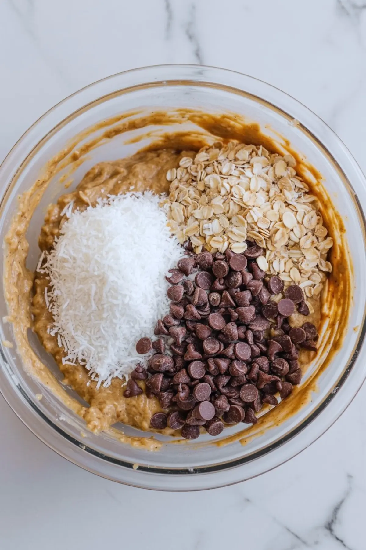 Overhead view of cookie dough topped with rolled oats, shredded coconut, and chocolate chips in a clear glass bowl.
