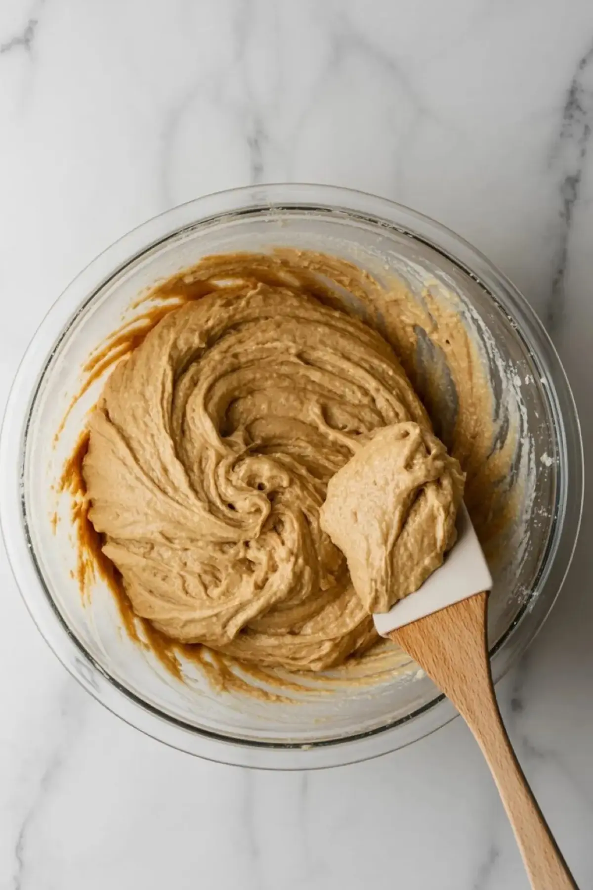 Glass bowl filled with thick paleo bread batter, partially mixed with a white silicone spatula on a white marble surface.
