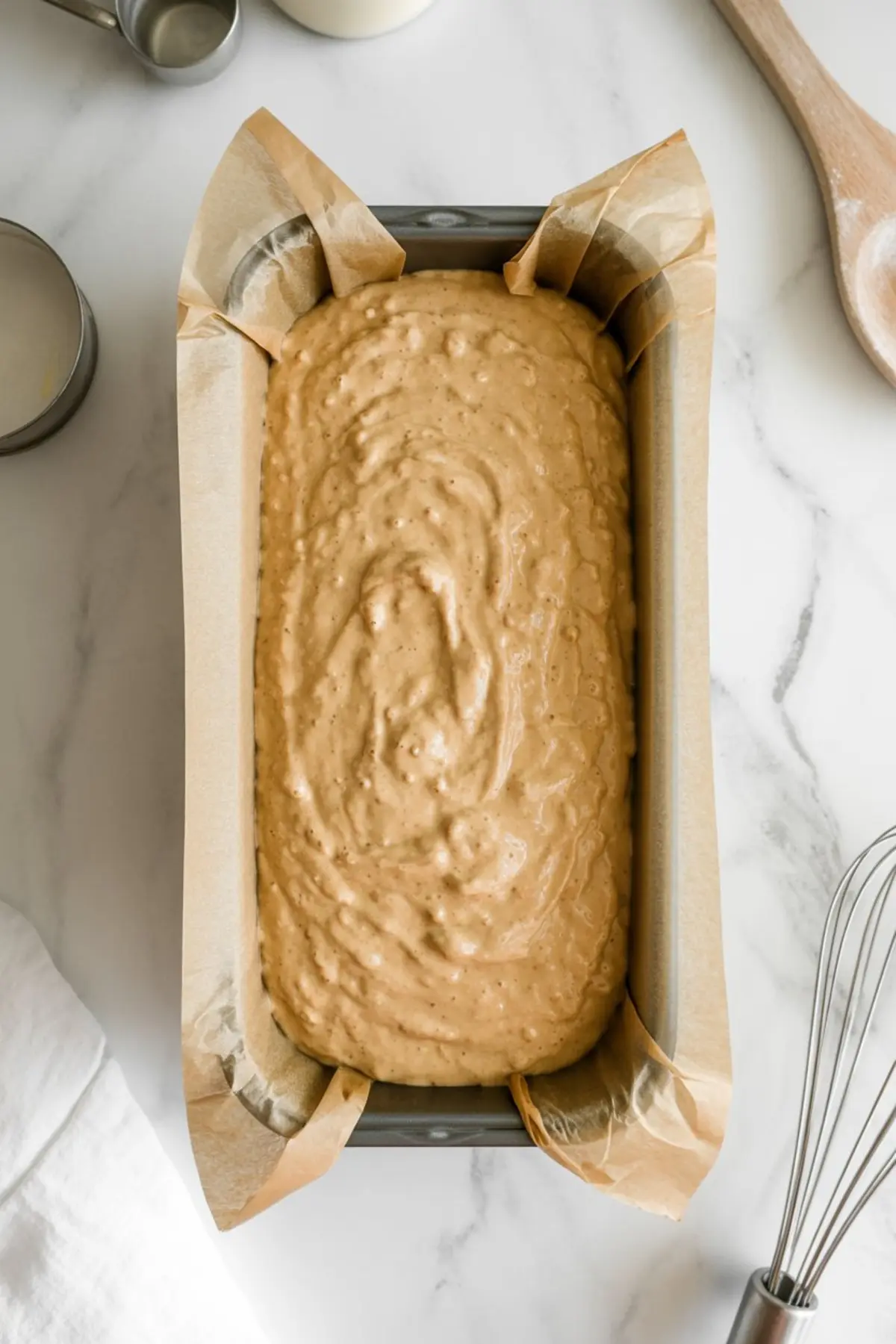 Unbaked paleo bread batter poured into a parchment-lined loaf pan, ready to go into the oven, set on a white countertop with baking tools nearby.

