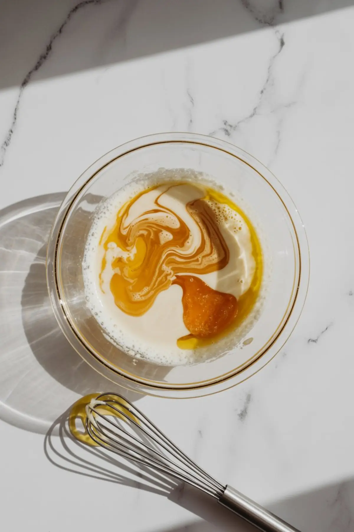 Top-down view of a glass bowl containing a mixture of almond milk, eggs, and golden sweetener, with a metal whisk nearby on a marble surface.
