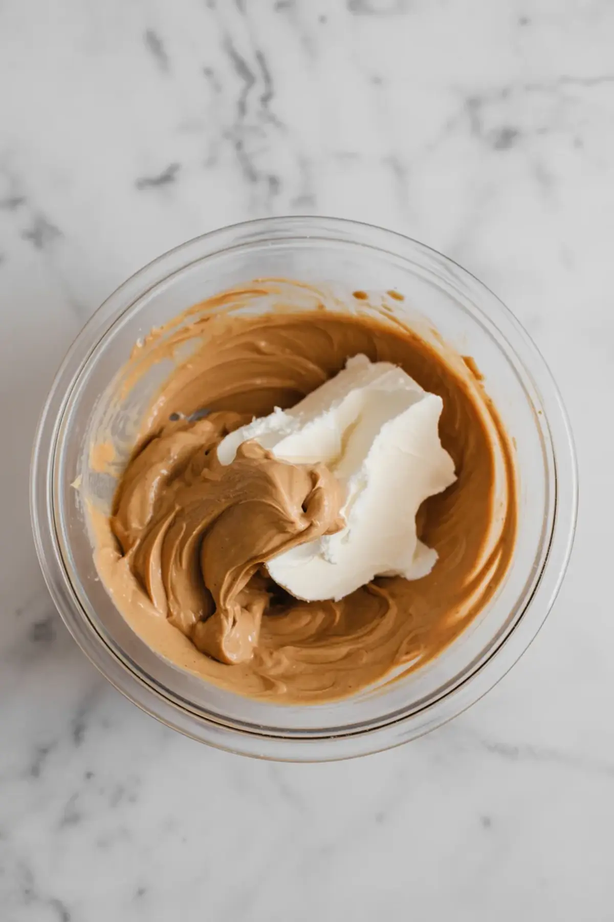 Glass mixing bowl containing partially combined cream cheese and smooth peanut butter for mousse preparation, on a white marble surface.