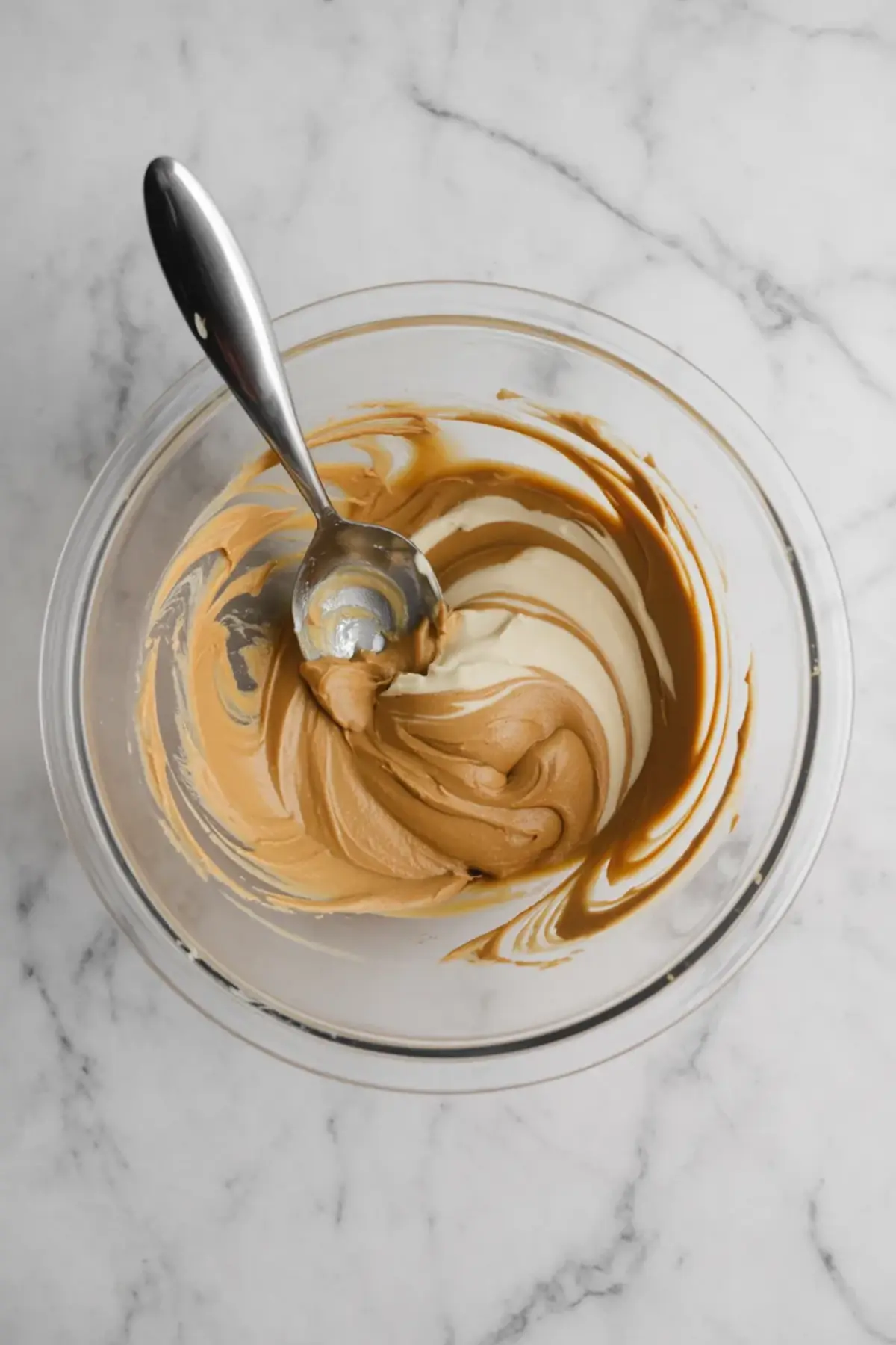 Partially mixed peanut butter mousse and whipped cream in a glass bowl with a spoon, showing the marbled blending stage on a white surface.