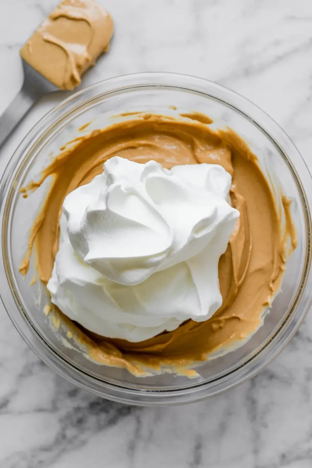 Large dollop of whipped topping added to a bowl of creamy peanut butter mixture, ready to be folded into mousse on a marble counter.