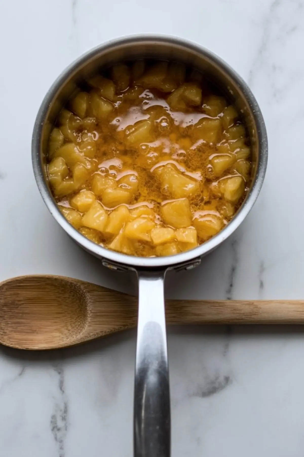 Cooked pineapple filling simmering in a small metal saucepan with visible pineapple chunks and glossy syrup, styled on a marble countertop with a wooden spoon beside the pan.
