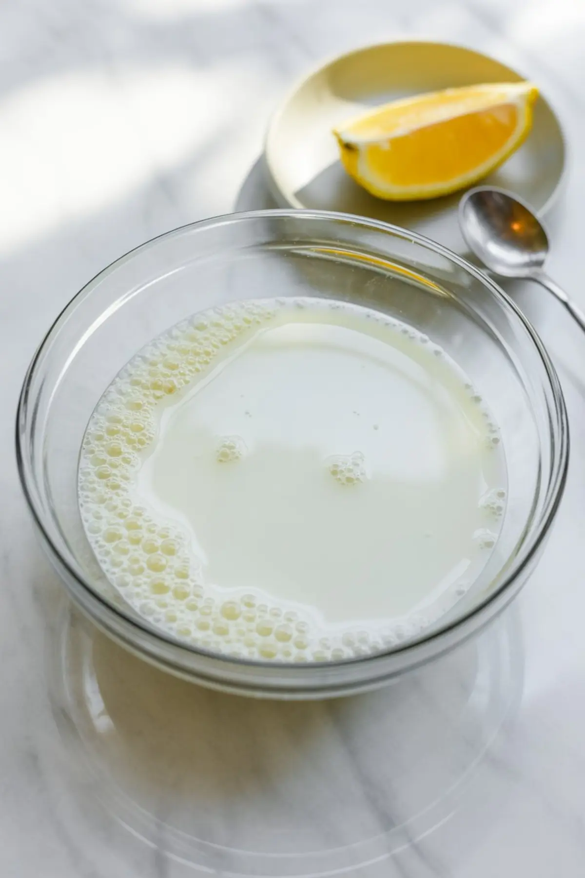 Glass bowl filled with milk on a marble surface, small bubbles visible on the liquid, lemon wedge on a plate and spoon nearby, baking ingredient preparation scene.