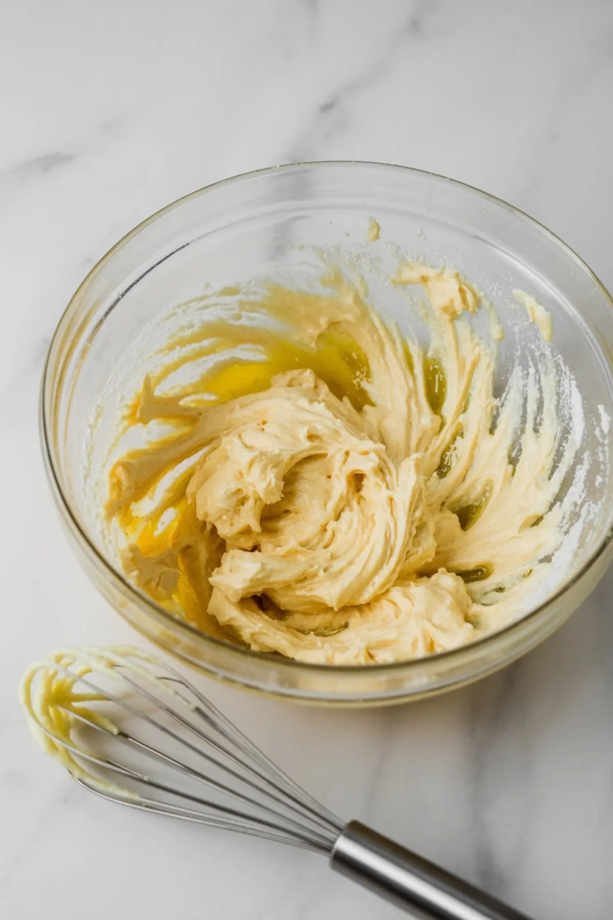 Glass bowl with thick cake batter being mixed, metal whisk coated in batter beside the bowl, creamy texture shown during mixing stage.