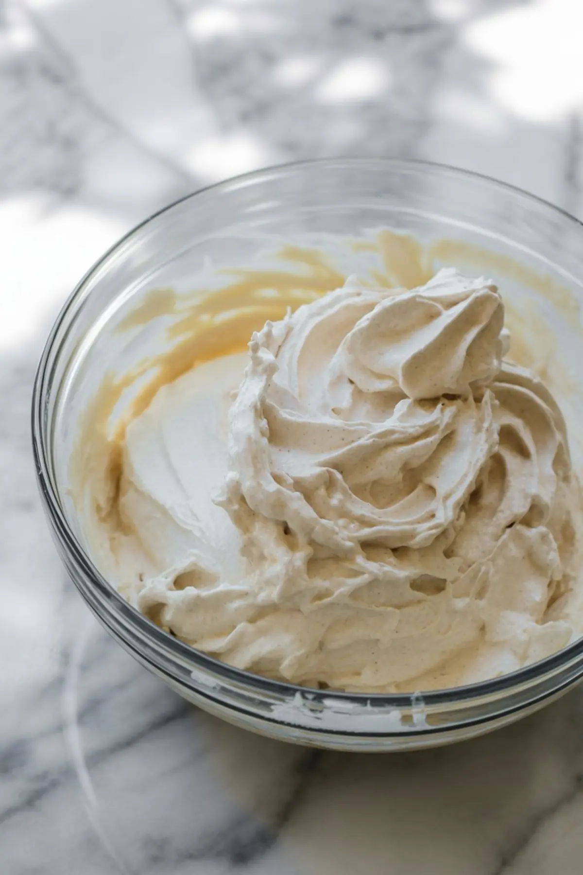 Glass bowl filled with whipped cream frosting, soft peaks and creamy texture visible, light natural lighting on marble countertop.