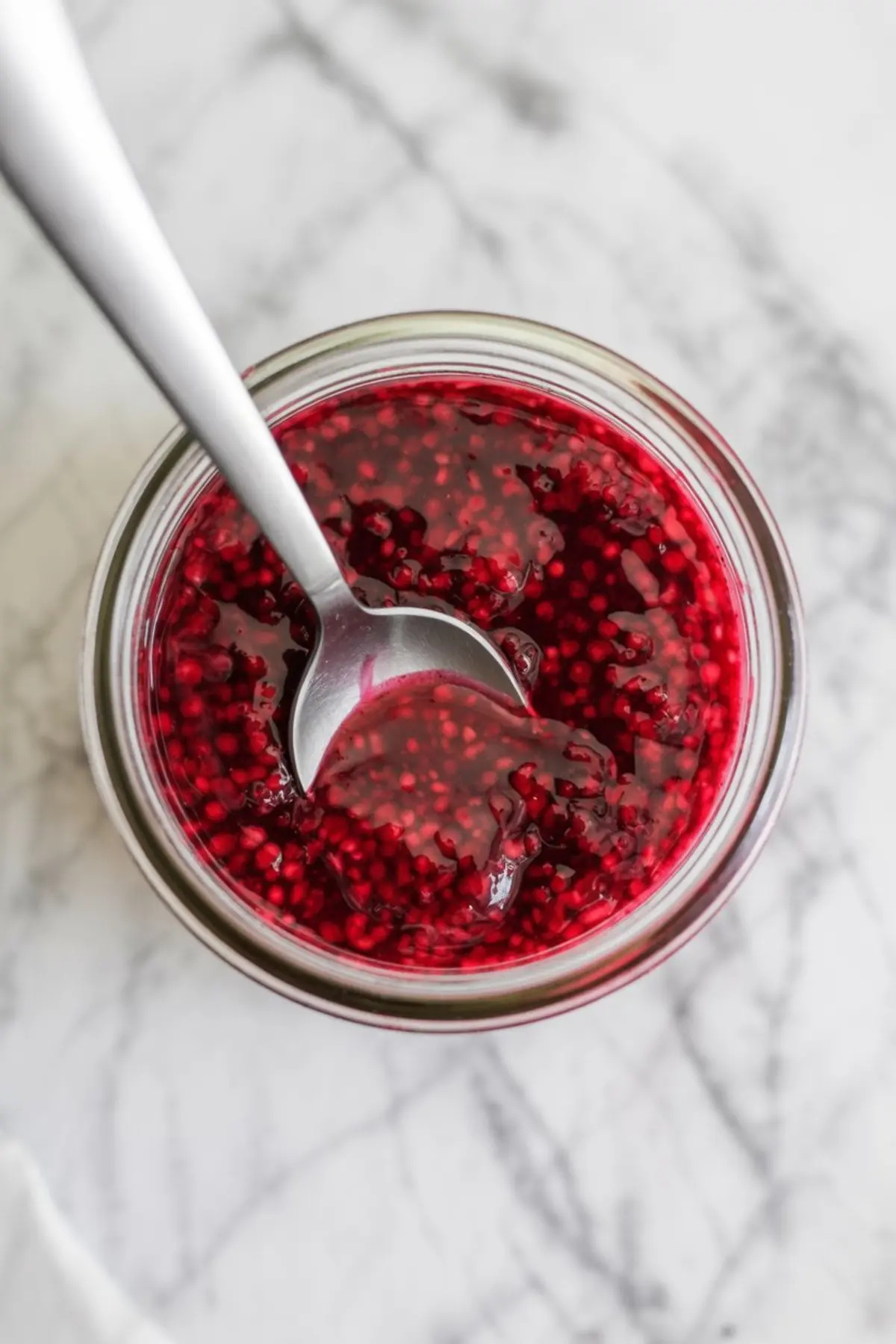 Glass jar filled with thick raspberry cake filling with visible seeds and a metal spoon, photographed from above on a light marble surface.
