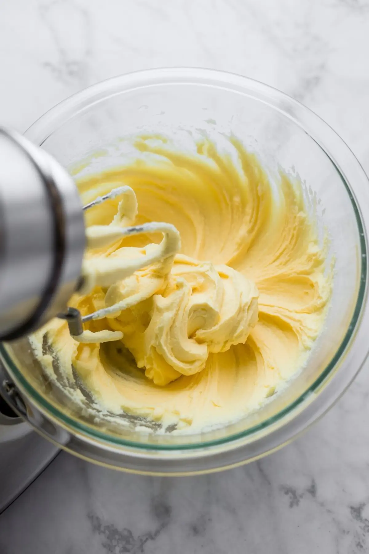 Electric mixer whipping creamy butter and sweetened condensed milk in a glass bowl, capturing the light yellow texture in the early stage of making Russian buttercream.
