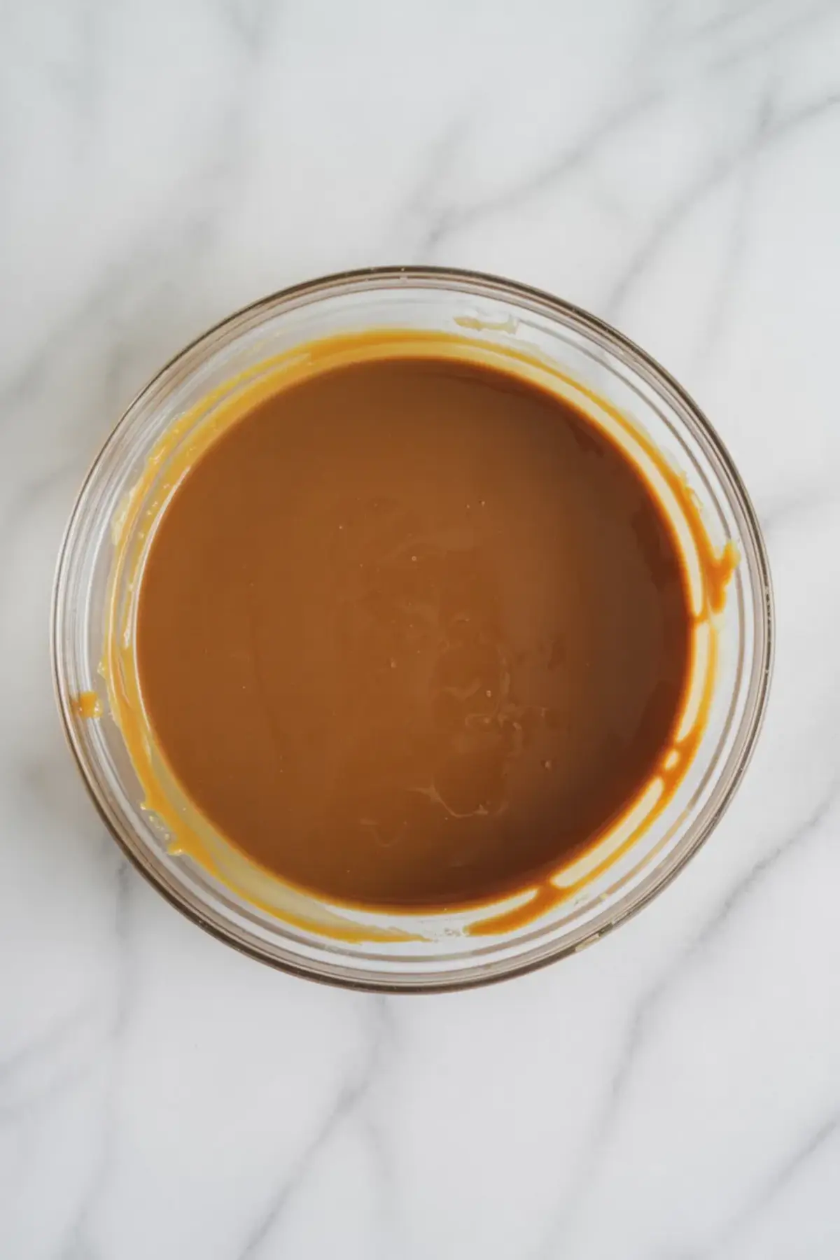 A clear glass bowl filled with smooth, golden-brown salted caramel sauce sits on a white marble surface, showing a glossy and thick consistency.
