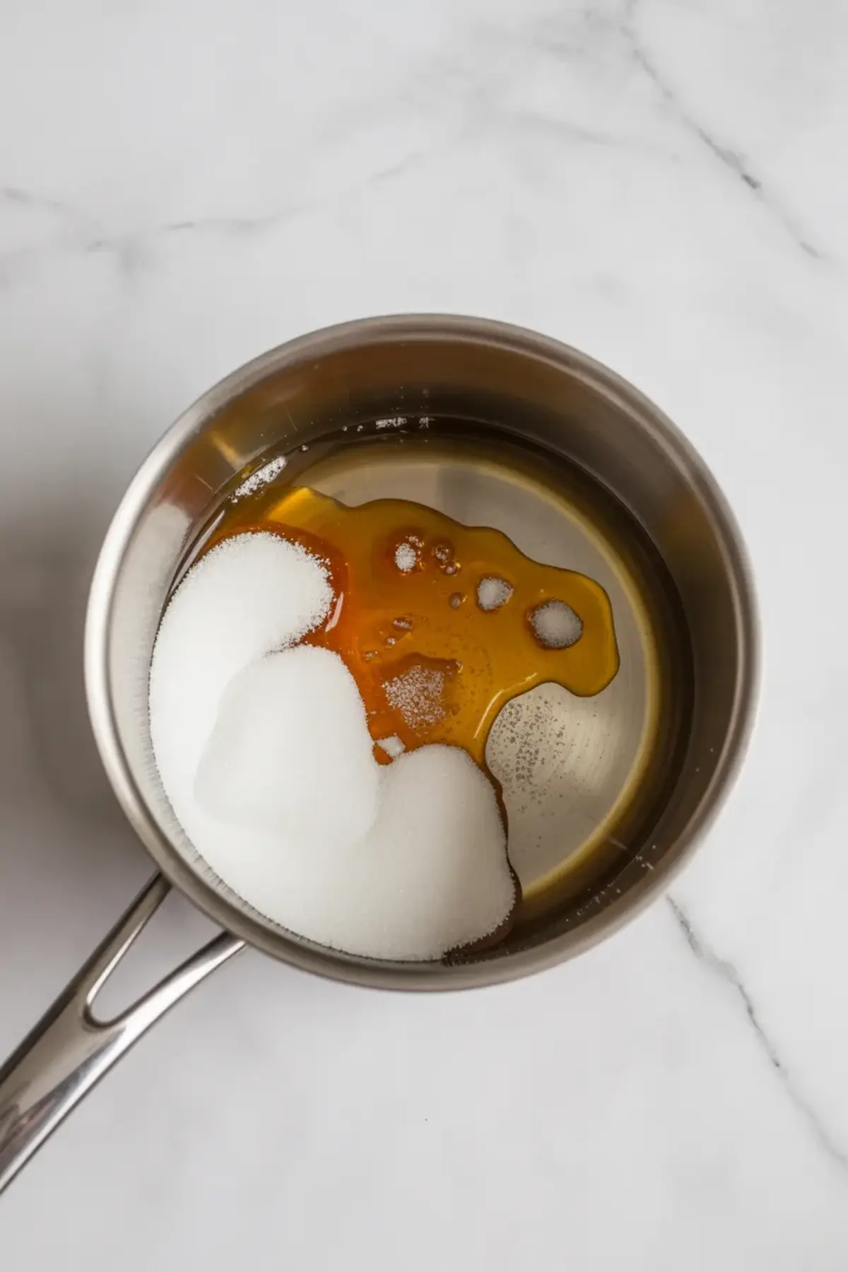 A stainless steel saucepan contains white granulated sugar and partially melted amber-colored sugar syrup, set on a white marble countertop during the early stage of caramel preparation.
